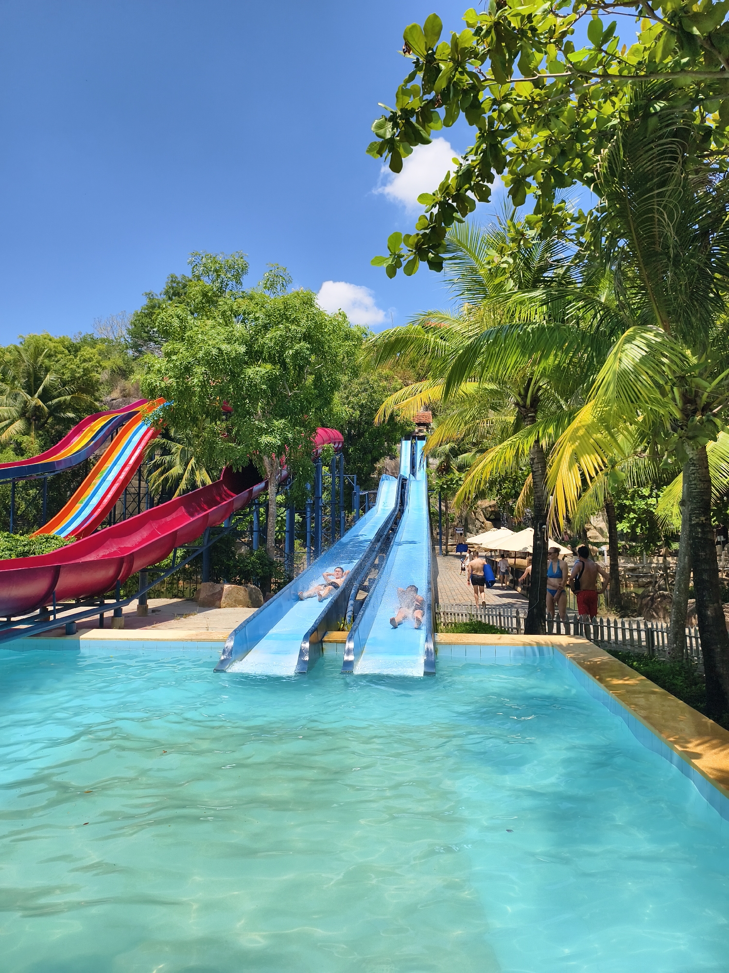 The image depicts a water park scene in Tay Nha Trang Ward, Vietnam. Two blue water slides terminate into a pool of light blue water. Two people are actively sliding down the blue slides. To the left, a red and multicolored slide is also visible. Palm trees and lush greenery surround the area. A small wooden fence separates the pool area from a walking path where several people are walking. The sky is bright blue with scattered white clouds.