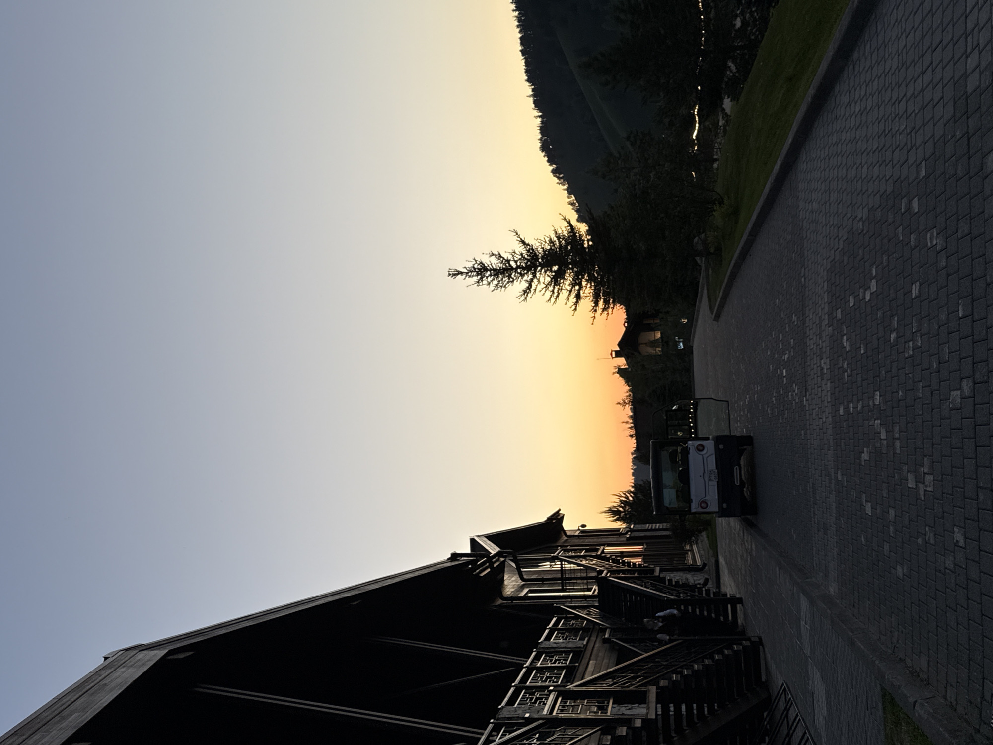 The image depicts an outdoor scene in Urlu-Aspak, Russia, at either dawn or dusk, characterized by a clear sky exhibiting a gradient from light blue at the zenith to a warm orange-yellow along the horizon.
On the left, a large, multi-story wooden lodge-style building with a dark, steeply sloped roof dominates the foreground. Its facade is constructed of dark wood, featuring multiple external staircases with elaborate wooden railings and balustrades. A single person, appearing to be an adult male in light clothing, is visible on one of these staircases on the building's lower left side. Several windows on the building reflect the warm light from the horizon.
In the mid-ground, a paved road made of dark grey interlocking pavers curves gently to the right, leading away from the building. A small, white, open-sided utility vehicle or golf cart is parked on this paved area, facing away from the viewer. The vehicle has a license plate on its rear, partially visible as "PT52 0677". The passenger-side door (from the viewer's perspective) is open.
To the right of the paved road, a grassy incline rises, dotted with several dark evergreen trees. A prominent, tall conifer stands silhouetted against the colorful sky in the middle-right. In the distant background, rolling hills covered with dark forest are visible, also largely silhouetted, with faint suggestions of lights or reflective surfaces near their base. The entire foreground, including the building, road, and immediate trees, is heavily shadowed, contrasting sharply with the bright sky and distant horizon.
