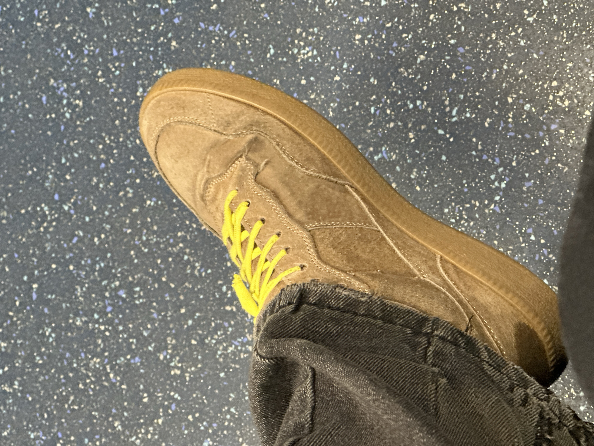 A top-down shot shows a person's foot clad in a brown suede-like sneaker with bright yellow laces. The sneaker is positioned atop a surface speckled with small, light-colored flecks on a darker background, giving the impression of a textured floor or ground. The person is wearing dark gray or black denim jeans with a cuffed hem. The scene provides no evidence of location in Moscow, Russia.