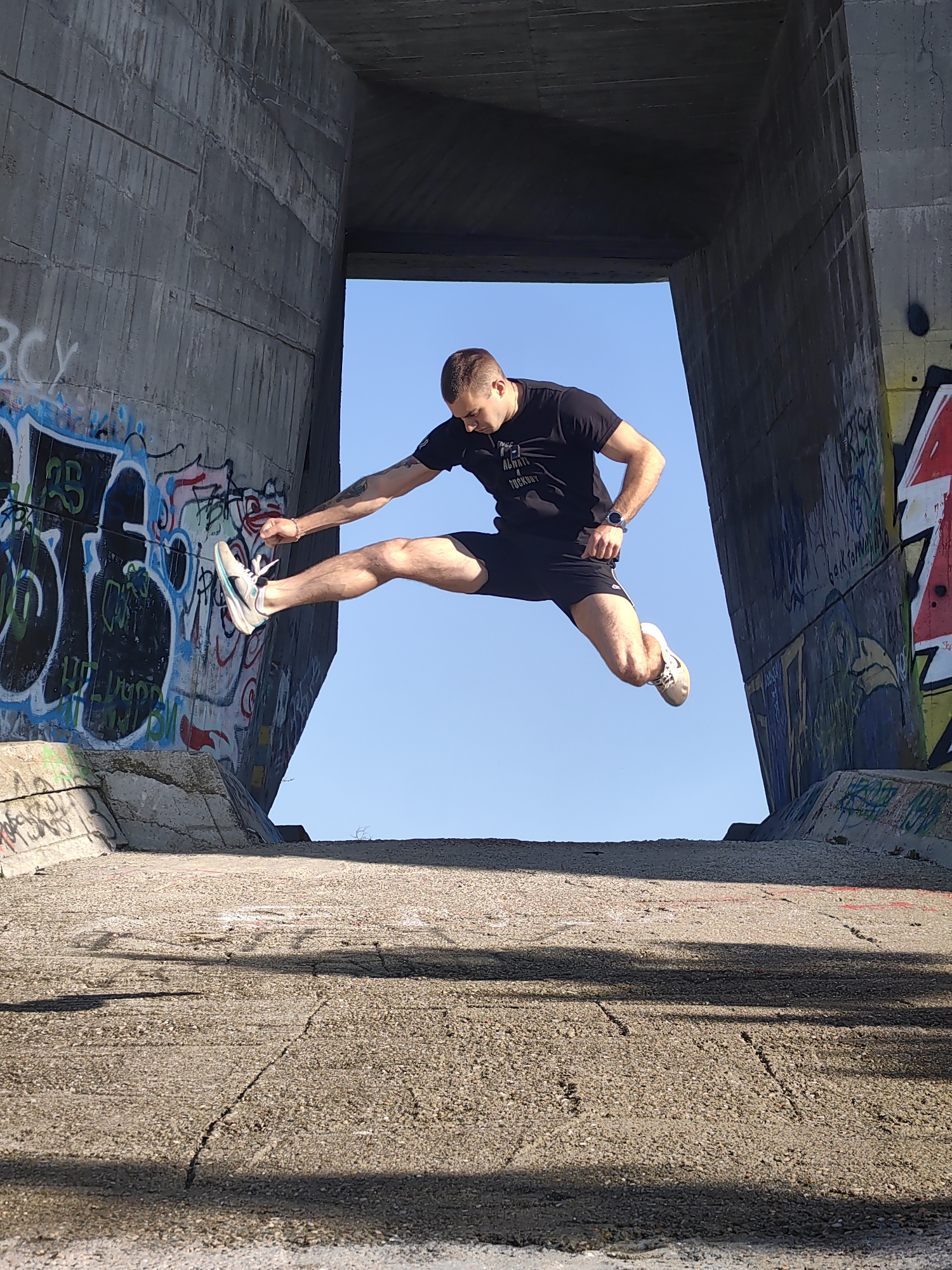 A man is captured mid-air in a dynamic jump between two concrete structures adorned with graffiti, set against a clear blue sky. The man is wearing a black t-shirt, black shorts, and sneakers. His left leg is extended forward, while his right leg is extended backward. His left arm is extended towards the left concrete structure. The location appears to be an underpass or an area with concrete architectural elements, possibly in Varna, Bulgaria, suggested by the graffiti style and the nature of the structures. The ground is concrete and has numerous cracks.