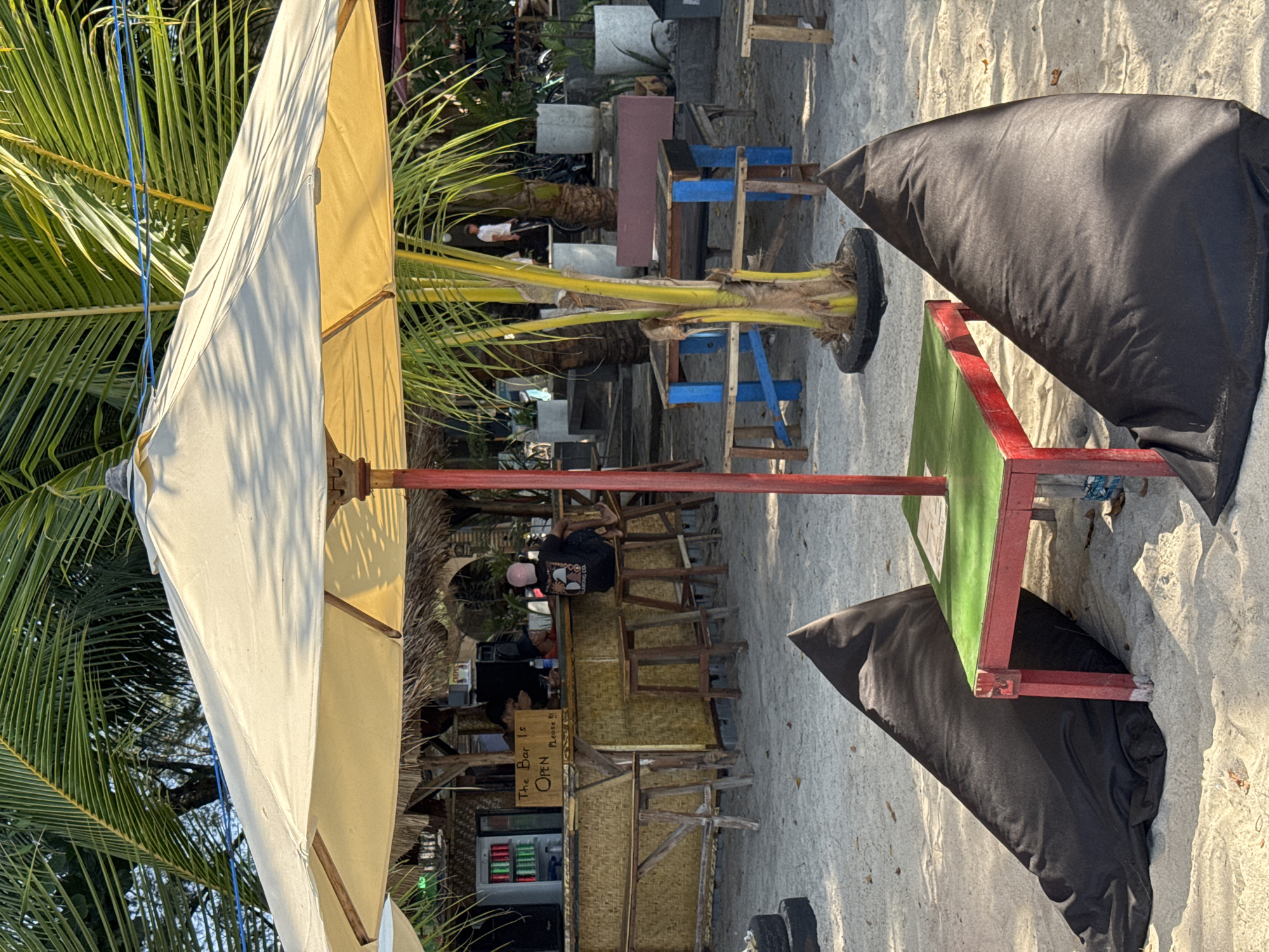 An outdoor beach bar in Unknown, Indonesia, features a thatched roof bar with drinks displayed on shelves and a sign stating "The Bar is OPEN Please!!!". Several wooden stools are positioned at the bar, and two individuals are visible behind it. In the foreground, a large beige umbrella shades a small red and green table accompanied by two large black beanbag chairs. Palm trees and other tropical foliage are present throughout the scene, with potted plants and a bicycle visible in the background. The ground is covered in sand.