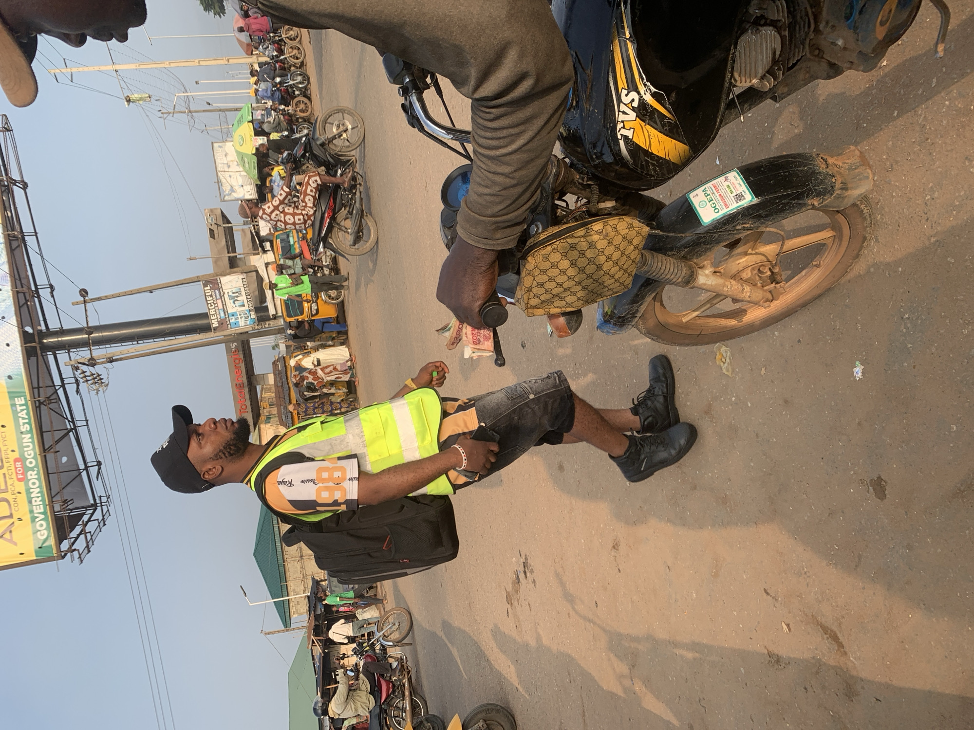 The image captures a street scene in Shagamu, Nigeria, with a focus on a man wearing a reflective vest and a black cap. He is standing, carrying a black backpack, and appears to be looking to his right. The background is bustling with activity, showing a road with various motorcycles and what appear to be rickshaws or small vehicles. Several people are visible, some on motorcycles and others walking. A large billboard with text including "GOVERNOR, OGUN STATE" is prominently displayed overhead. A motorcycle in the foreground, to the right of the man, has a sticker on its fender that reads "OGEPA EMISSION PERMIT". The ground is dusty, characteristic of a road in this region. The lighting suggests it is daytime, with shadows cast on the ground.