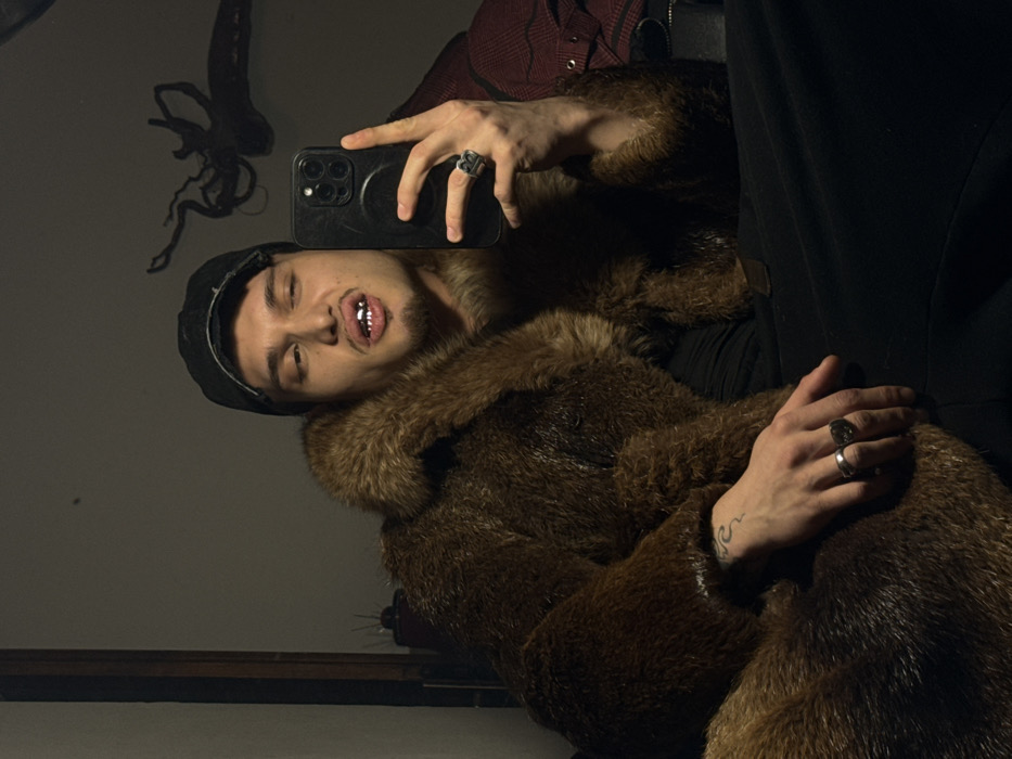 A male individual is depicted indoors, capturing a selfie with a black smartphone held in his right hand. He wears a dark, distressed baseball cap and a voluminous brown fur coat. His upper teeth are fitted with silver-colored dental grillz, and he has a dark goatee. His left eye is partially closed, while his mouth is slightly open. The subject's left hand, displaying two rings and a wave-pattern tattoo on the forearm, rests on the fur garment. A large, dark ring is visible on his right ring finger. The background is dimly lit, featuring a dark vertical frame on the left and a dark, amorphous wall decoration resembling an octopus or tangled branches on the upper right. A red, patterned fabric is partially visible on the far right. The scene is located in Sofia, Bulgaria.