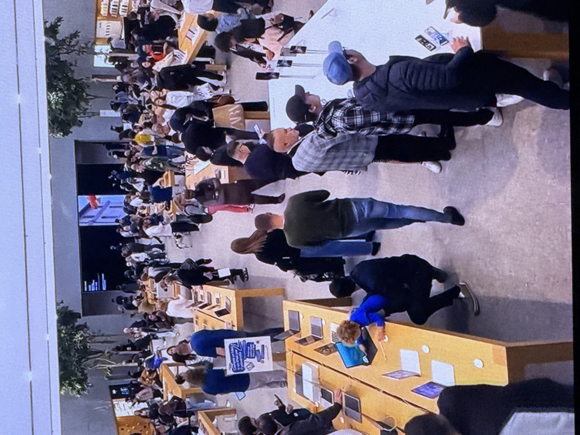 This media file captures a scene from a modern retail environment, likely an electronics store. A diverse group of people are seen interacting with various electronic devices displayed on wooden tables. The subjects are a mix of adults, appearing to be shoppers and possibly staff, engaged in browsing, testing, or purchasing products.
The objects prominently featured are numerous laptops and tablets, laid out on the tables for public access. The tables themselves are a warm, light wood color, creating a contemporary and inviting atmosphere. The overall setting is an indoor space with bright, diffused lighting, suggesting it's daytime, though no specific time of day can be definitively determined. The presence of lush green plants in the background, particularly on the left side of the frame, hints at a well-designed retail space, possibly a flagship store.
The activity taking place is primarily shopping and product interaction. People are seen leaning over tables, pointing at screens, and holding devices. Some individuals appear to be in conversation, suggesting a lively commercial atmosphere. There's a sense of engagement and curiosity from the shoppers.
Notable details include the variety of clothing worn by the people, indicating a casual to semi-casual dress code. The floor appears to be a neutral, light-colored material, possibly concrete or tile, contributing to the modern aesthetic. No specific weather conditions are discernible as the scene is indoors.
There is some visible text in the image. A white sign with blue lettering is held up by one of the individuals, though the text itself is not clearly legible. Another sign, possibly an advertisement or product information, is visible on one of the tables.
Considering the provided location context of Dubai, United Arab Emirates, the scene is consistent with the modern retail experiences found in large shopping centers or dedicated brand stores within the city. The upscale appearance of the store and the diverse crowd are common characteristics of retail spaces in Dubai.