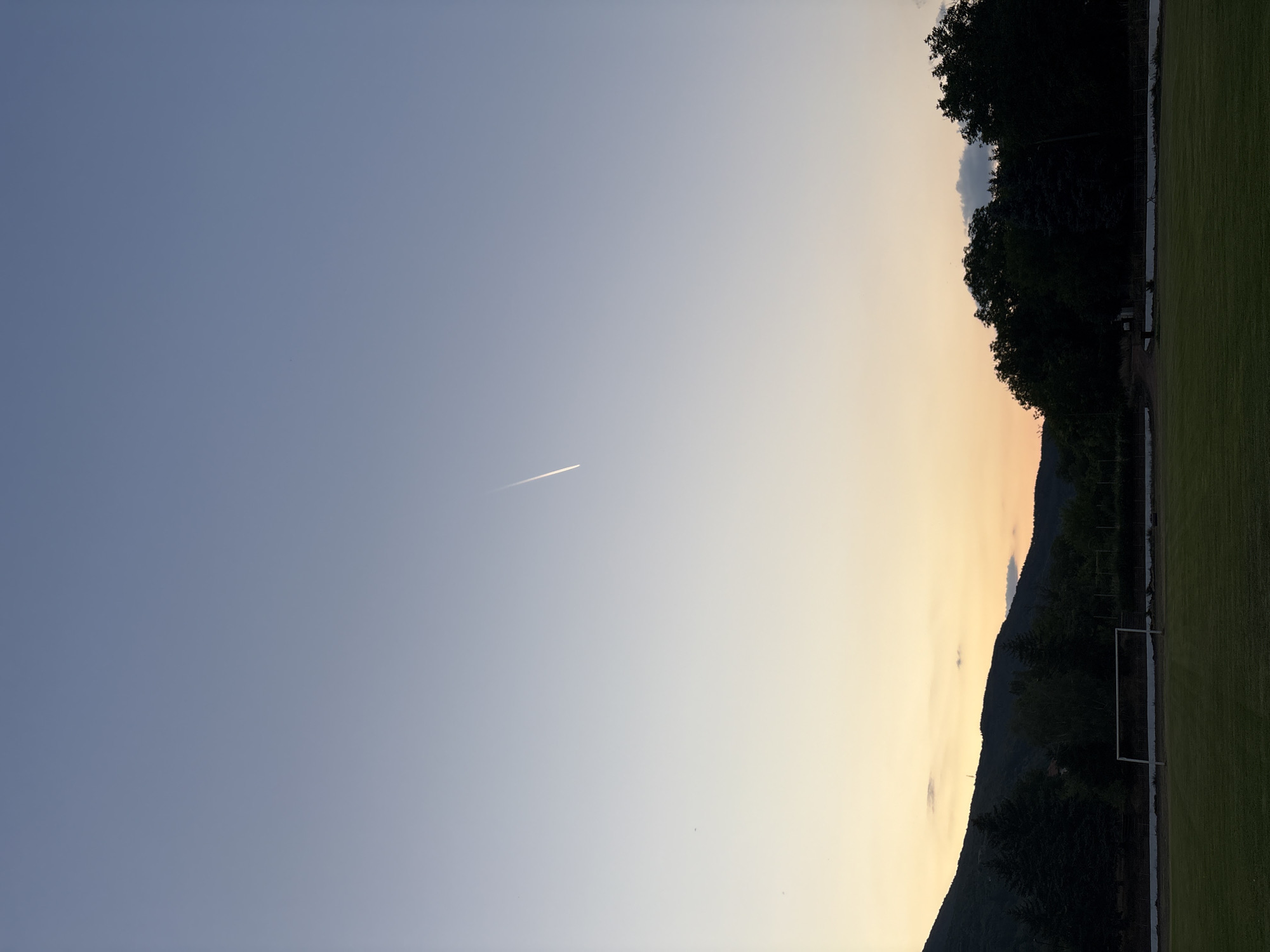 This is a vertical, outdoor, daytime photograph taken in Boboshevo, Bulgaria. The image captures a clear sky transitioning from a pale blue at the top to a soft yellow and orange hue near the horizon. A thin, white contrail from an aircraft is visible diagonally across the upper portion of the sky. Below the sky, a dark silhouette of trees and a mountain range lines the horizon. In the foreground, a green grassy field, possibly a sports field, is visible with a white soccer goal at the far end. The lighting suggests either early morning or late evening. No people are present in the frame.