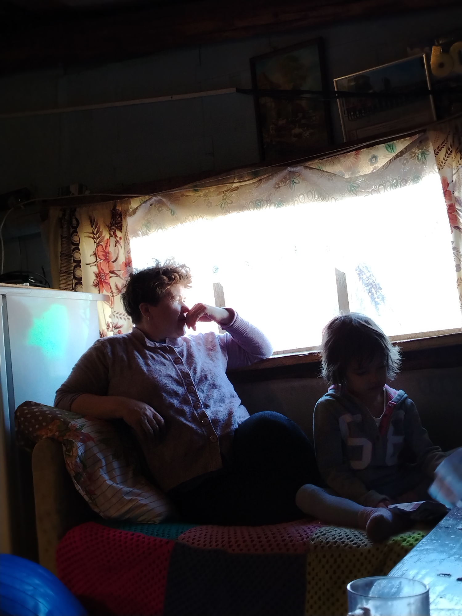 In a dimly lit room in Гожан, Russia, a woman and a young girl are seated on a sofa. The woman, with short curly hair, is wearing a lavender button-down shirt and dark pants. She is leaning against the window frame, her hand near her face, seemingly lost in thought. Beside her sits the young girl, dressed in a hooded sweatshirt, her attention focused downwards, perhaps on an object in her lap. The window behind them is bright, covered by floral curtains. Adjacent to the woman, to the left, is a white refrigerator. The sofa is covered in a patterned fabric. In the upper right corner, two framed pictures are visible on a shelf. A blue object is partially visible at the bottom left, while a glass sits on a table at the bottom right.