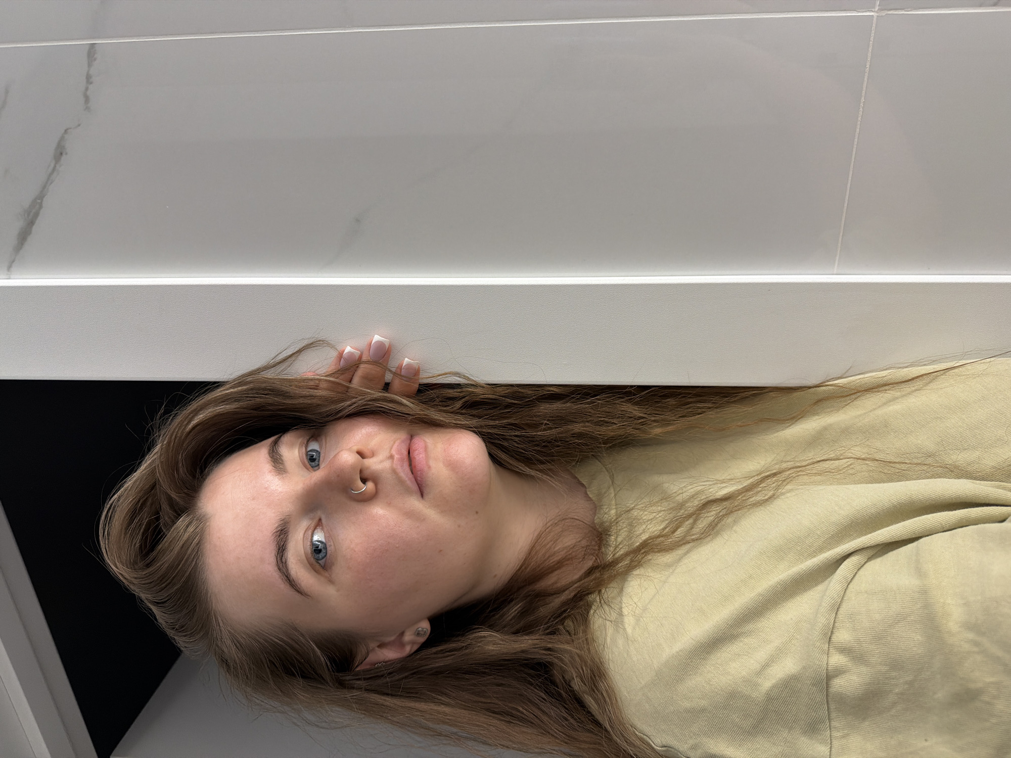 A young, fair-skinned woman with long brown hair and blue eyes looks directly at the camera. She has a nose ring and a small earring. Her nails are painted with a white French tip. She wears a light greenish-yellow shirt. She is positioned close to a white shelf or ledge, with white tiled walls above. The location is unidentifiable as Chaikovsky, Russia, from the provided image.