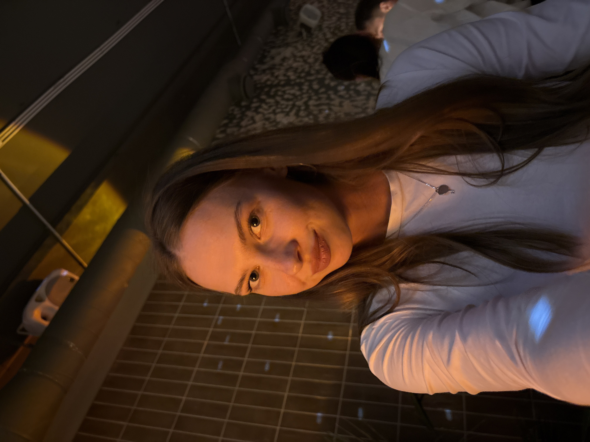 A young woman with long, light brown hair, wearing a white long-sleeved shirt and a silver necklace with a key-shaped pendant, looks directly at the camera. She has light skin and is wearing subtle makeup. Her right hand is visible, extended towards the camera. The background shows a tiled wall to the left and a textured wall with white dots to the right. In the upper left corner, a white speaker is mounted on a pipe. The scene appears to be indoors, possibly a bar or restaurant, with warm lighting casting shadows and reflections on the woman's shirt. The presence of other people in the background suggests a public space. The image does not provide specific visual cues to pinpoint a location within the city of Perm, Russia.
