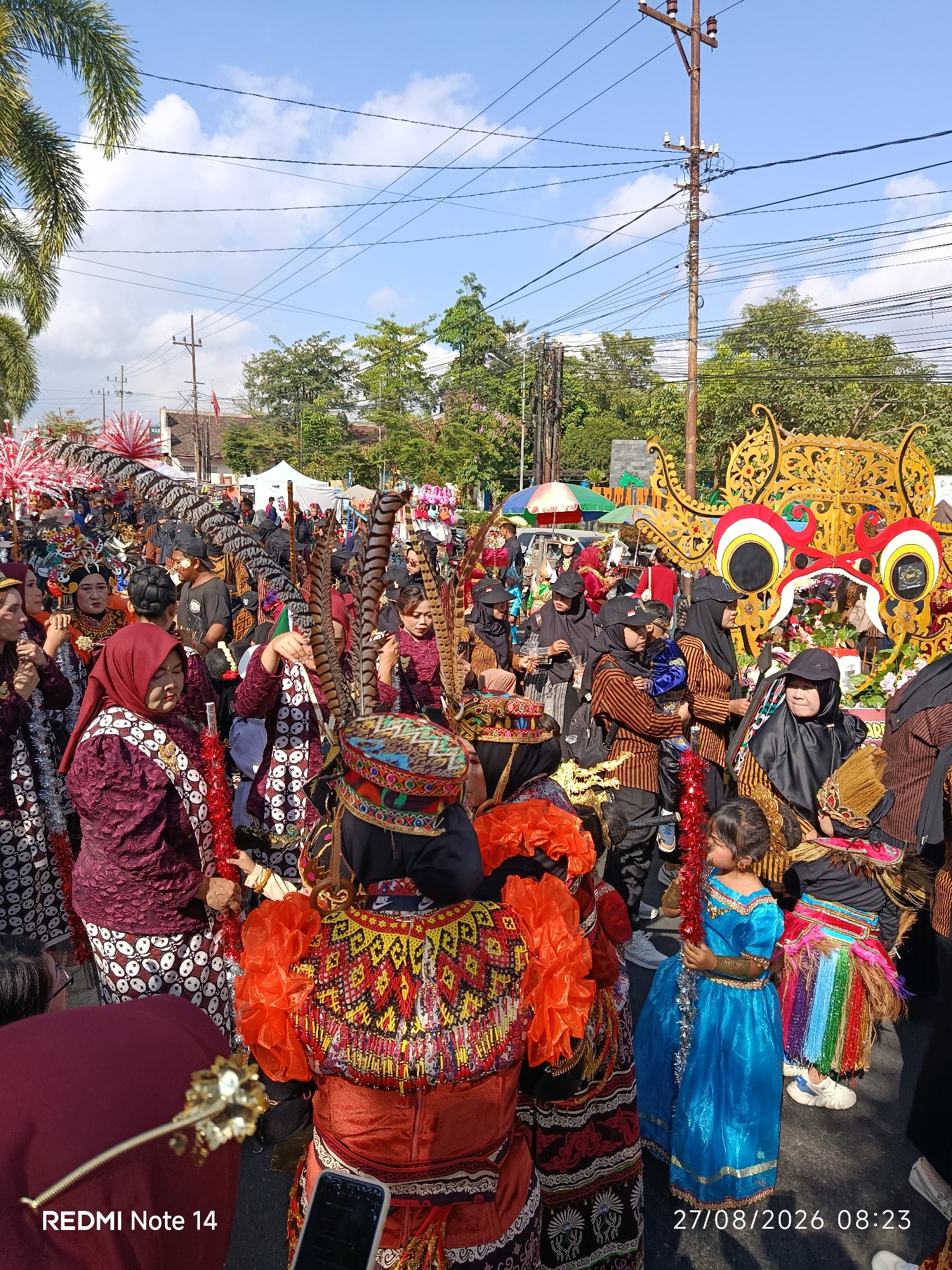The image captures a vibrant street scene in Kalangbret, Indonesia, likely during a cultural procession or festival. A large crowd of people, many adorned in traditional attire, fills the street. The clothing is rich with detail, featuring intricate patterns, colorful fabrics, and elaborate headdresses adorned with feathers. Several individuals wear dark headscarves, and others are dressed in striped shirts. A child in a bright blue dress stands out amidst the crowd, holding a red tinsel decoration. In the background, a large, ornate golden structure, possibly part of a float or display, is visible, with striking red and white eye motifs. Tents and stalls suggest a market or event setup along the street. The environment is bright and sunny, with power lines crisscrossing overhead and lush green trees lining the background. The presence of a smartphone in the foreground, displaying the text "REDMI Note 14" and a date and time stamp "27/08/2026 08:23," indicates the image was captured using a mobile device and provides a contemporary timestamp for the event.