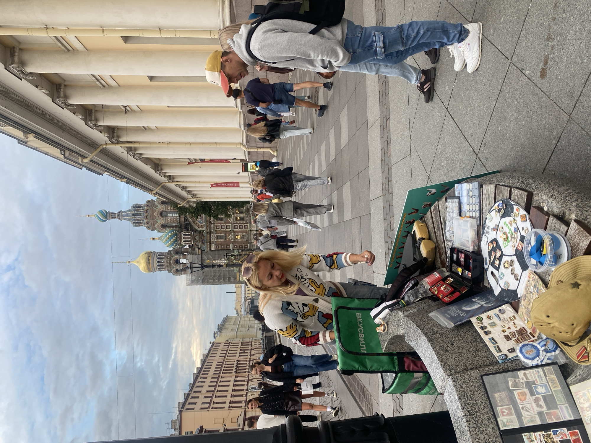 This is a street-level photograph taken on a cloudy day in Saint Petersburg, Russia, likely in the vicinity of the Church of the Savior on Spilled Blood, as evidenced by its distinctive onion domes visible in the background. The foreground is dominated by a low, curved, stone-and-wood bench, which serves as a makeshift display area for various items. A woman with blonde hair, wearing a cardigan with cartoon characters, is leaning over the bench, appearing to be arranging or selling items. Scattered on the bench are collections of stamps, pins, badges, and a wide-brimmed hat adorned with medals. A bright green insulated delivery bag is positioned next to the bench.
Several people are walking on the paved sidewalk in the midground and background. To the right, a man wearing a cap and a grey hoodie, and a woman in blue jeans are walking away from the camera. Further back, a diverse group of pedestrians, some carrying shopping bags, are moving along the sidewalk. The architecture on the right side of the image features a building with a colonnade of large, light-colored columns and a pale yellow facade. The overall scene suggests a lively urban environment with a mix of tourists and locals.