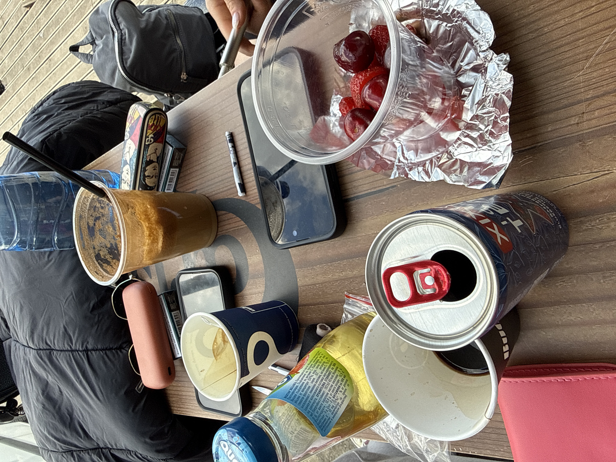 The image depicts a cluttered table setting, seemingly outdoors, with an array of personal items and consumables. There's a mix of opened and partially consumed beverages, including a coffee in a plastic cup with a straw, an open beer can, and an opened bottle of what appears to be juice. A transparent container holds strawberries and cherries, some of which are wrapped in aluminum foil. Personal items visible on the table include two smartphones, a pink AirPods case, a cigarette case, a pen, a blue and white patterned paper cup, a pink wallet, a bottle of water, and a gray backpack. The table has a wooden surface with a circular design. Based on the OCR content from the juice bottle (Пастьоризиран. НЕКТАР ЯБЪЛКА), it indicates that the juice is Bulgarian, which strongly suggests that the location of the image is likely in Bulgaria. Without additional context clues, it is impossible to pinpoint the specific location within Sofia, Bulgaria. The image does not show any people.