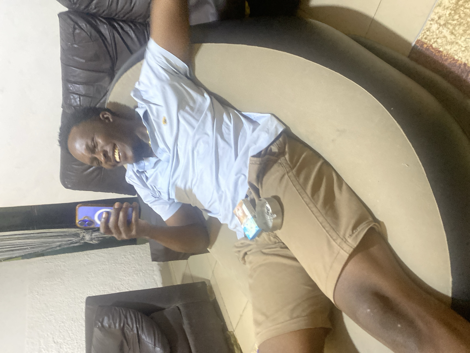 This media file features a young man relaxing on a sofa in an indoor setting, likely a living room, located in Kubwa, Nigeria. The image is rotated 90 degrees, showing the man lying on his back with his head to the left of the frame.
The subject is a young man with dark skin and short, dark hair. He is dressed in a light blue short-sleeved collared shirt, featuring a small, golden emblem on the left chest, and tan/khaki shorts. A gold-colored piece of jewelry is visible around his neck. He is actively engaged with a smartphone, which he holds in his left hand. The phone has a distinctive blue case, and its screen displays a prominent blue circular icon with a white symbol.
The man's expression is one of evident joy and amusement; he is laughing heartily, with his mouth open and teeth visible. His body language suggests relaxation, with his right arm bent and raised towards his head. On his right thigh, a small, blue and white packaged item, possibly a pack of gum or similar, is visible.
The setting includes a sofa with dark brown/black armrests and back, contrasting with a lighter beige-colored seat portion. Parts of a light-colored wall and a textured rug or carpet are visible in the background. The lighting appears to be artificial, typical of an indoor environment. No specific brand names or readable text are discernible on the packaged item or the phone screen.