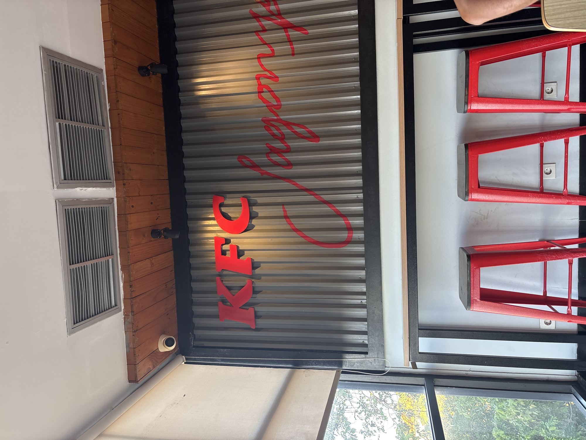 The image shows the interior of a KFC restaurant in Kuta Selatan, Indonesia. A corrugated metal wall features prominent red "KFC" lettering and a cursive red script that reads "Lagoon". Above the wall, a wooden panel runs horizontally, with two ventilation grilles on the white ceiling. A security camera is mounted on the left side of the wooden panel. In the foreground, three red bar stools are positioned in front of a counter. A window on the left side of the image looks out onto green foliage.