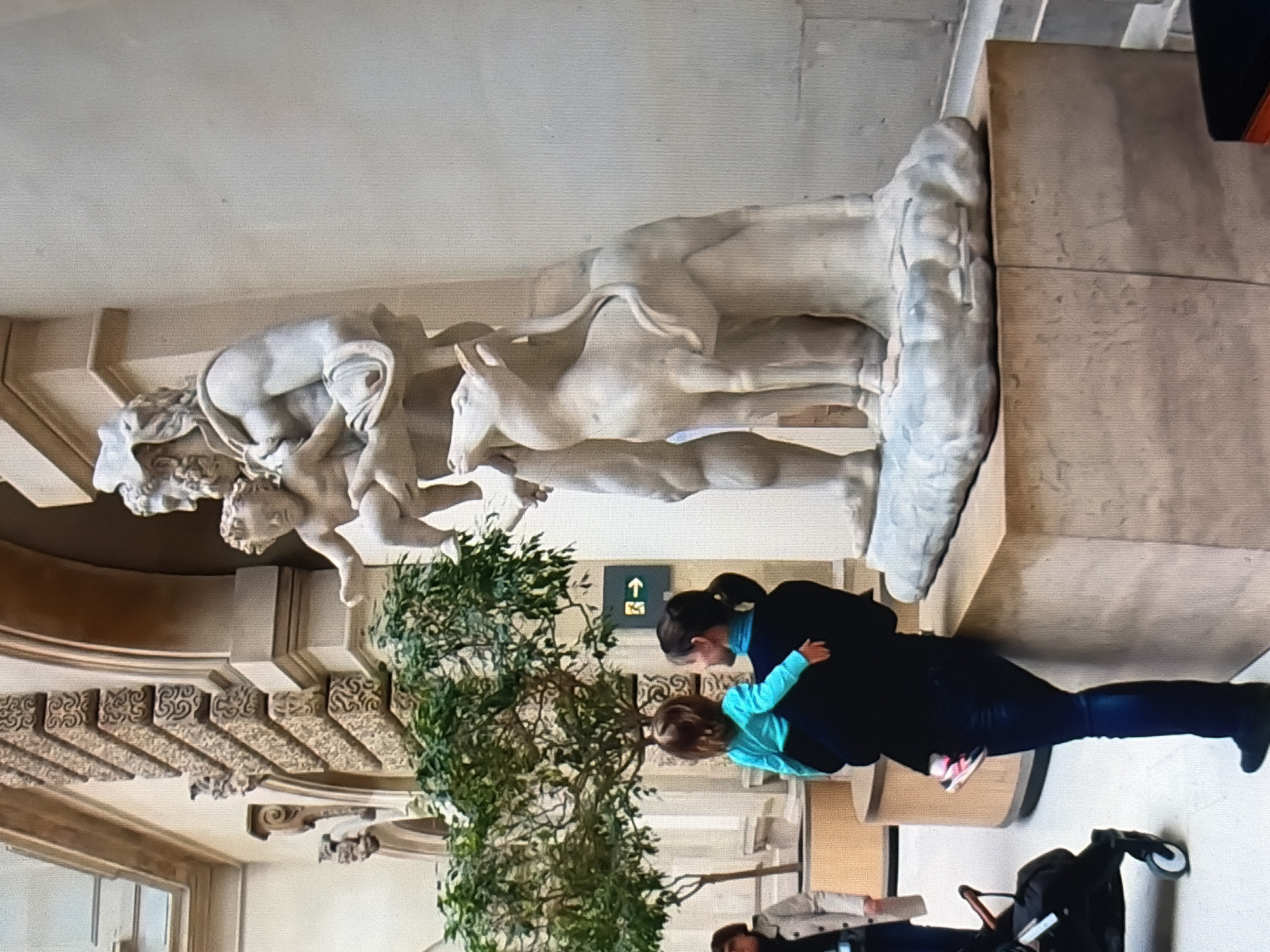 A full shot captures a classical marble sculpture of a male figure holding a child and a goat, positioned in a museum or art gallery. The sculpture is on a light brown pedestal. In front of the sculpture, a woman is holding a toddler. The woman is wearing a dark jacket and dark pants, and the toddler is wearing a bright blue long-sleeved shirt and pink and white shoes. To the left of the woman, a stroller is visible. In the background, an indoor tree with green leaves and architectural elements of the building are visible. A directional sign with an arrow pointing right is mounted on the wall behind the tree. The lighting appears to be natural, coming from a large window or skylight.