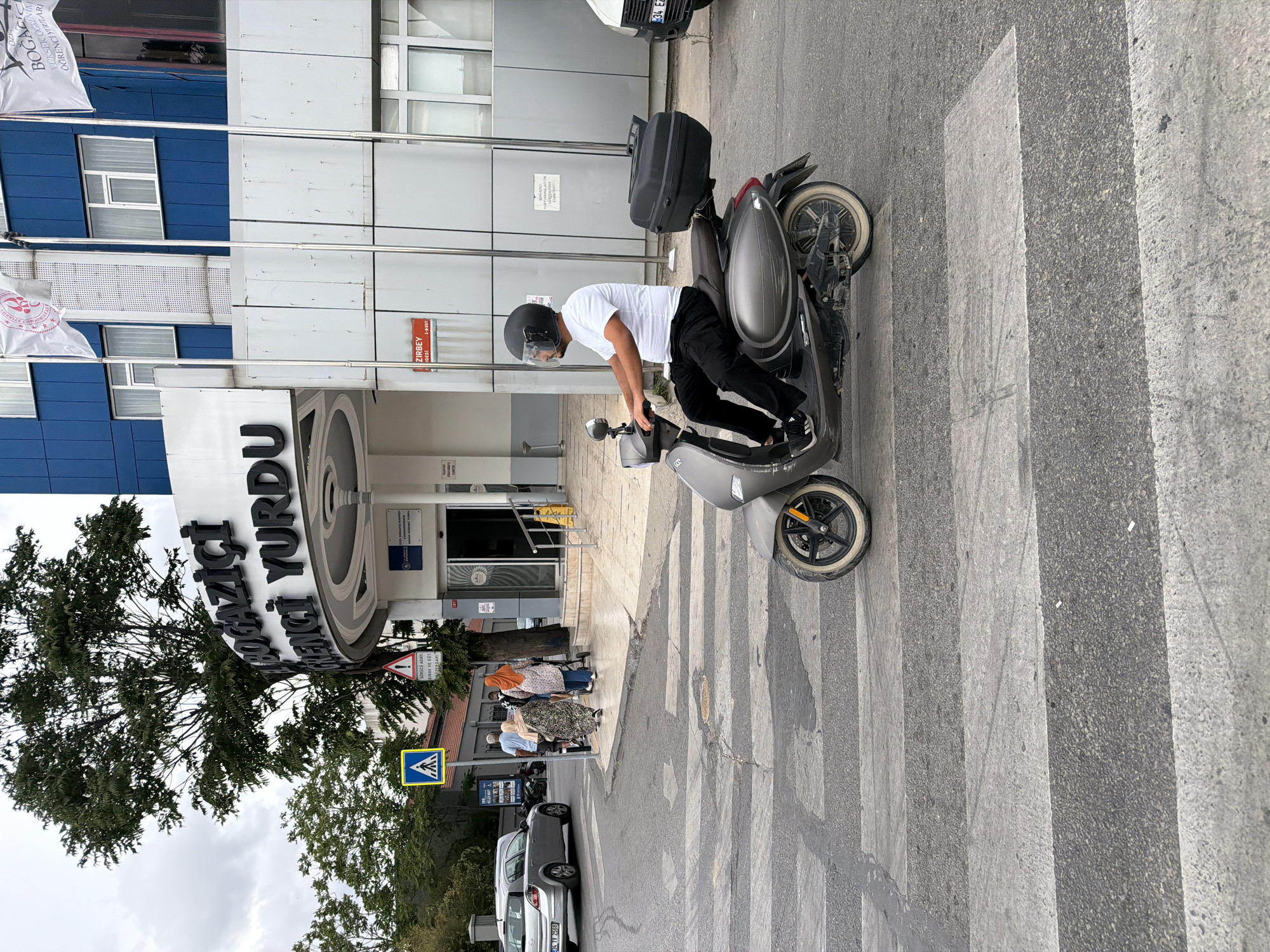 A man wearing a white t-shirt, black pants, and a black helmet rides a grey scooter across a pedestrian crossing. The scooter has a black storage box mounted on the rear. In the background, a building with blue and white siding is visible, with flags flying in front. A sign on the building reads "BOĞAZİÇİ ÖĞRENCİ YURDU" which translates to "Boğaziçi Student Dormitory". To the left of the building, a silver car is parked. Further left, a pedestrian crossing sign is displayed. A group of people, two women and one man, are walking on the sidewalk in front of the building. The scene appears to be in Kadıköy, Turkey.