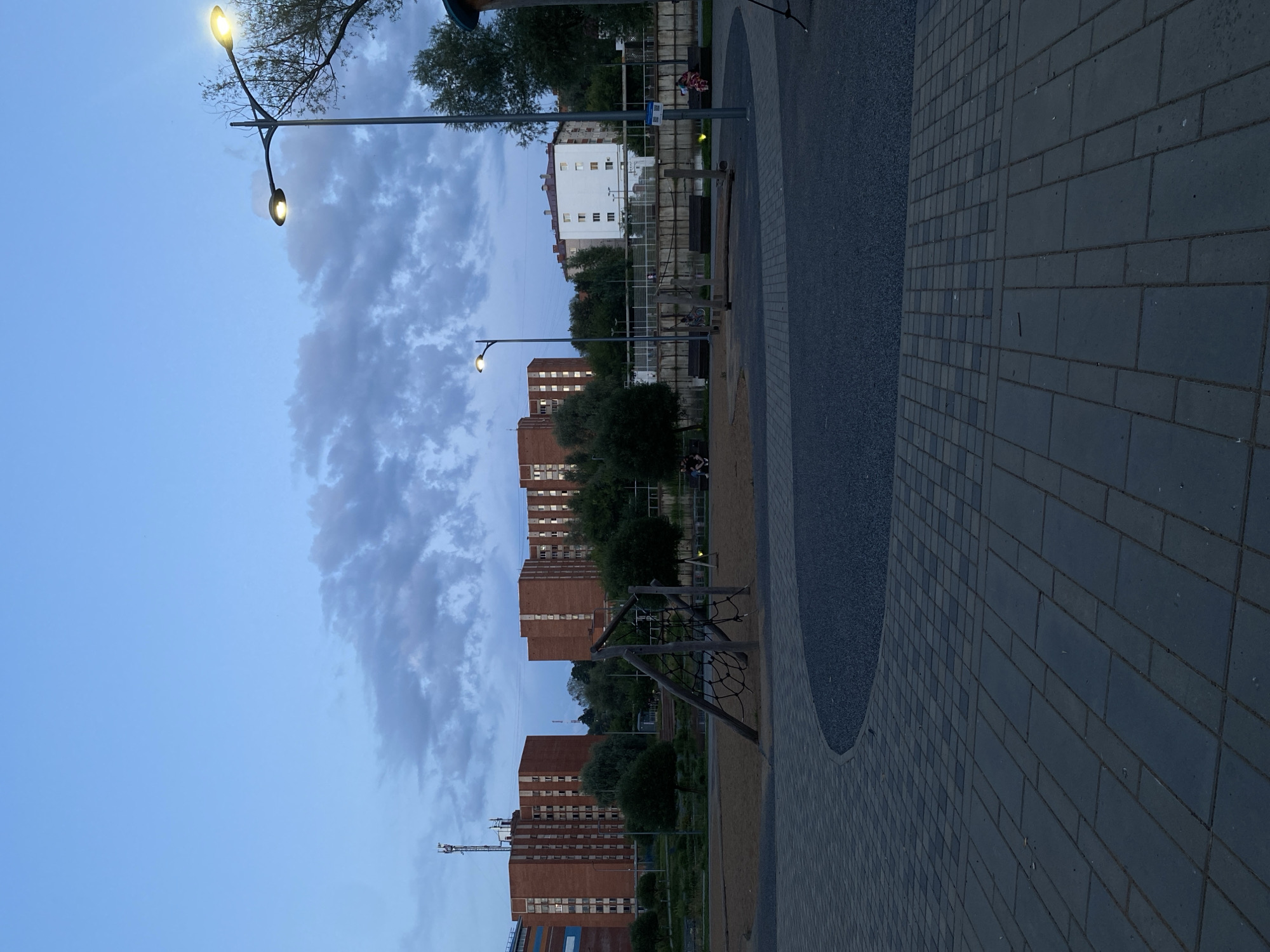 The image depicts an outdoor scene in Votkinsk, Russia, during twilight. In the foreground, a paved area with two distinct patterns of dark grey paving stones forms a curved pathway. Beyond this, a patch of dark gravel is visible. In the midground, a children's playground with a sand surface features wooden climbing structures. Benches are placed along a paved walkway behind the playground, and several trees and bushes provide greenery. In the background, multiple multi-story brick apartment buildings stand under a cloudy, pale blue sky. Two streetlights are illuminated, casting a warm glow. A cell tower is visible in the distance. Two individuals are seated on benches, one of whom appears to be interacting with a child on a bicycle. Another person is sitting on a bench with a child. The overall atmosphere is calm and suggests a residential area at the end of the day.