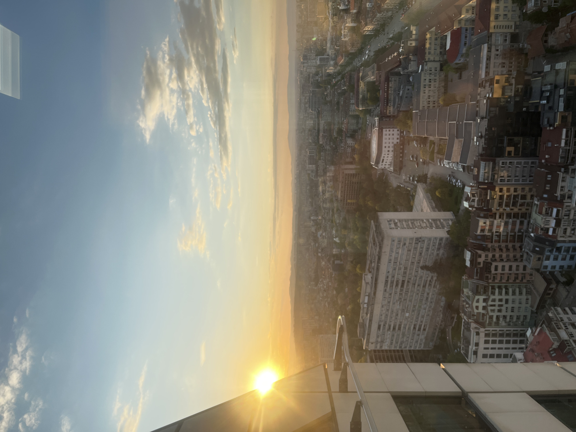 A panoramic view of the cityscape of Sofia, Bulgaria, is captured at what appears to be sunset or sunrise, casting a warm, golden glow across the urban landscape. The sky, transitioning from a pale blue overhead to vibrant orange and yellow hues at the horizon, is dotted with scattered white clouds, illuminated by the low-lying sun.
The sun, a bright and intense orb, is positioned low on the left side of the frame, partially obscured by the foreground structure and creating a pronounced lens flare. Below the horizon, the dense city sprawls into the distance, featuring a mix of buildings of various architectural styles and heights, along with visible streets and pockets of green spaces. Faint outlines of mountains can be seen on the distant horizon, adding to the expansive vista.
In the immediate foreground, the top edge of a modern building, likely a balcony or observation deck, is visible, with its grey panels and reflective surfaces. No people or visible text are discernible in the photograph. The atmosphere is somewhat hazy, characteristic of golden hour light.