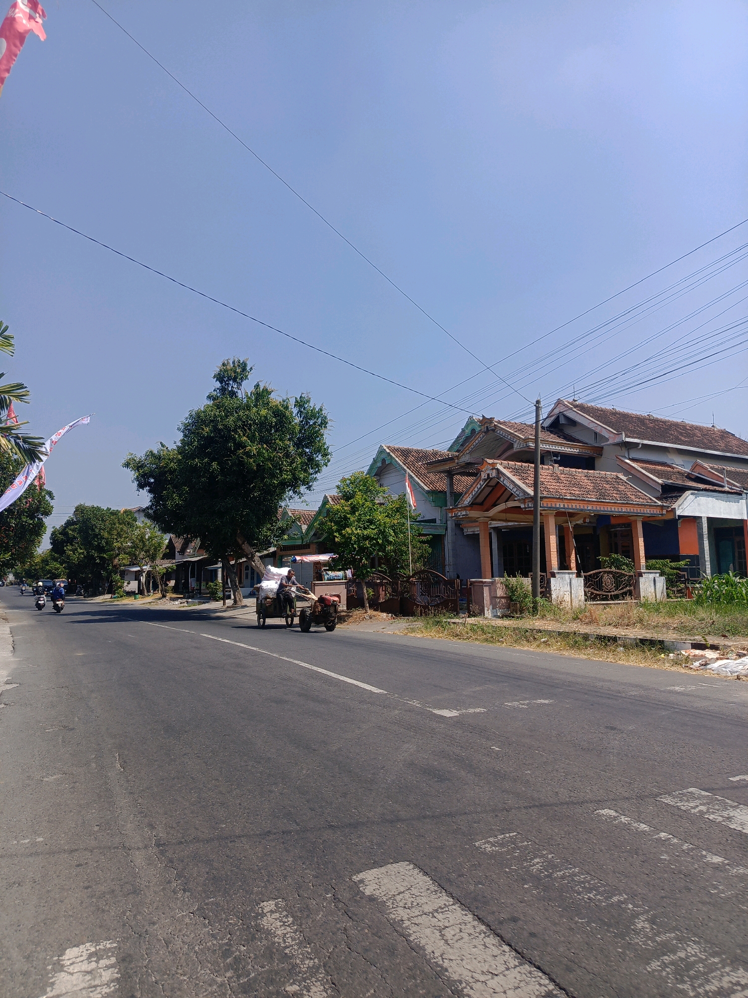 The image depicts a scene in Sukowinangun, Indonesia, on a sunny day with a clear blue sky. A paved road runs through the frame, with a zebra crossing visible in the foreground. On the right side of the road, a row of houses with tiled roofs are visible, interspersed with greenery. The houses appear to be single-story or two-story structures with various architectural styles. A large tree with dense foliage stands prominently in front of some of the houses. On the left side of the road, more trees line the street, and a few motorcycles are seen in the distance. A small, two-wheeled cart loaded with white sacks is being pulled by a man on a small motorized vehicle, moving along the road. The cart appears to be a utility vehicle used for transport. The overall impression is a rural or semi-urban environment with residential buildings and daily life activities taking place on the road.