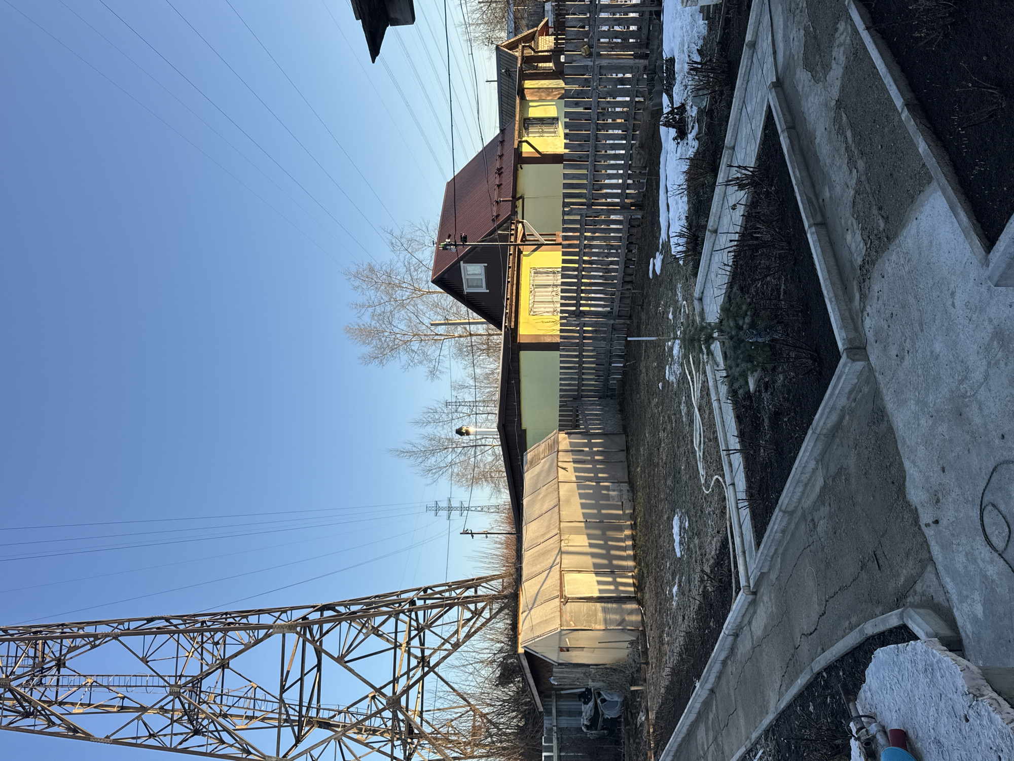 The image depicts a scene in what is likely a residential or suburban area, situated in Chaikovsky, Russia. The environment appears to be a sunny, early spring day, with patches of snow still visible on the ground.
A prominent feature of the landscape is a large electrical transmission tower, constructed of metal lattice, positioned to the left of the frame. Overhead, high-voltage power lines stretch across the clear blue sky.
Adjacent to the tower, a greenhouse-like structure with translucent walls stands, casting long shadows on the ground.
In the middle ground, there is a yellow, possibly wooden, house, partially obscured by a wooden fence. The house has a rust-colored roof and visible windows.
To the right, there are concrete garden beds, with some soil or planting material visible inside them. A hose can be seen stretched across the grass.