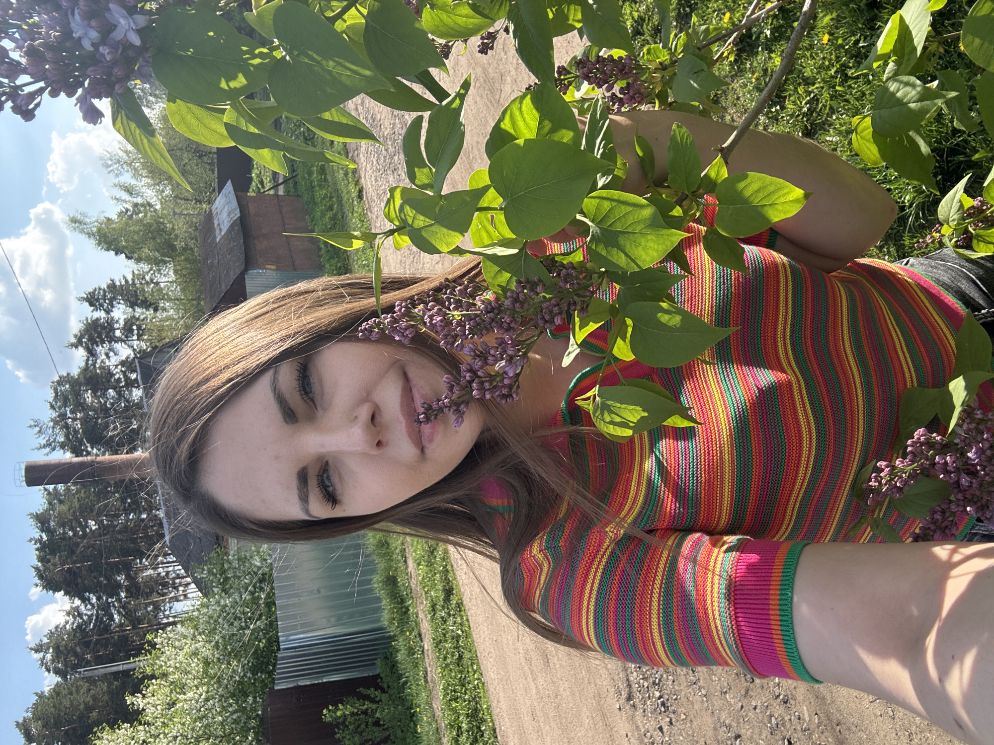 The image shows a young woman outdoors, posing with lilac flowers. She wears a brightly striped, long-sleeved shirt. The background reveals a rural or suburban setting, likely in Раздолье, Russia, with simple buildings, fences, trees, and a dirt road. A tall chimney or pipe is visible in the distance behind the trees on the left. Sunlight dapples the scene.