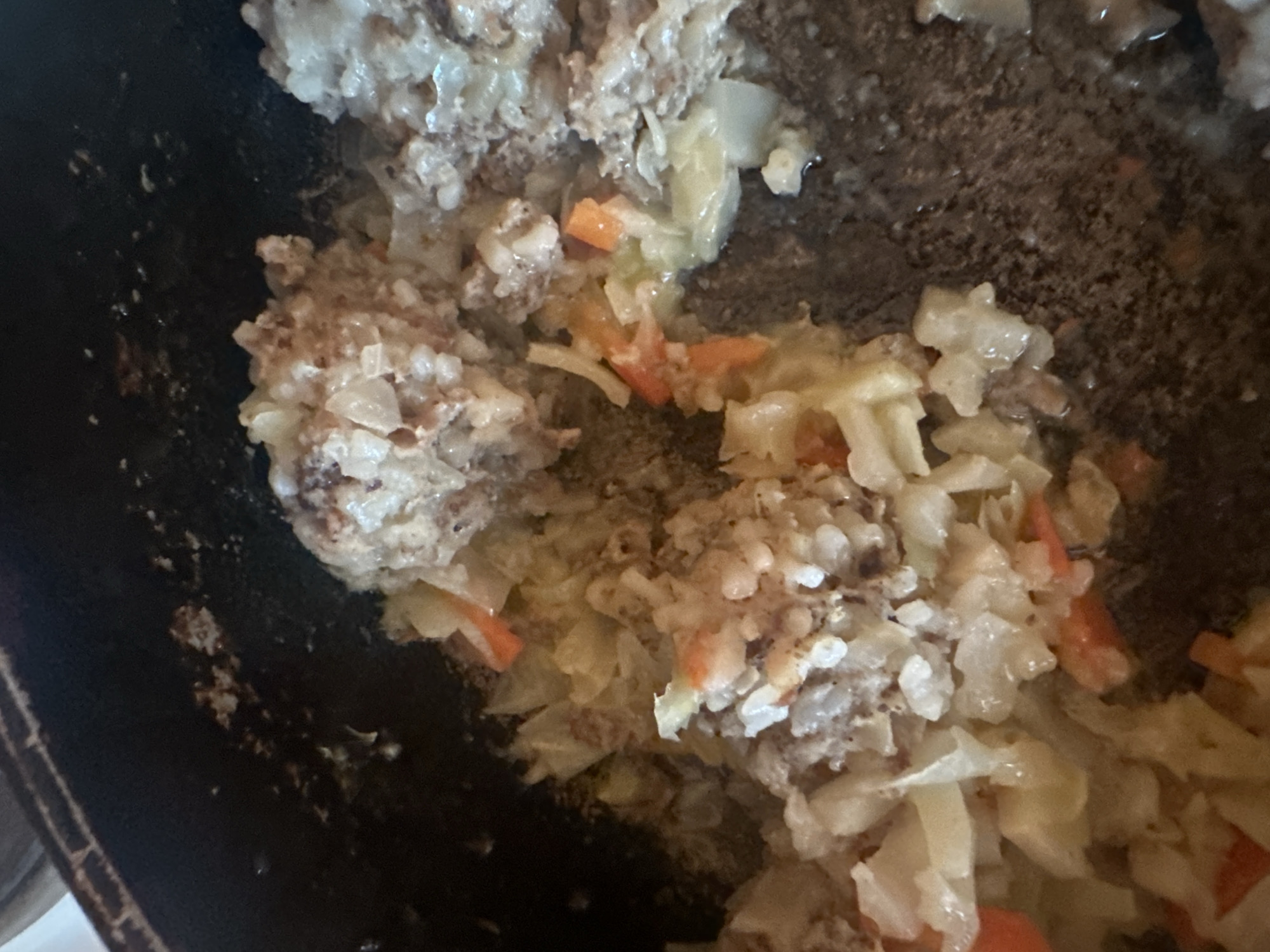 A close-up, high-angle shot captures a mixture of food ingredients cooking in a dark, possibly cast-iron, vessel. The dish appears to be a hearty preparation, possibly a stew or a filling for another meal.
The primary subjects are the food ingredients: sizable chunks of brownish-gray ground meat, mixed with white, soft-looking grains of rice. Interspersed throughout the mixture are translucent-white pieces of chopped cabbage and bright orange diced carrots. The overall texture is moist and somewhat lumpy, indicating the ingredients have been cooked together. Some liquid is visible among the food, contributing to its moist appearance.
The cooking vessel itself is dark, likely metal, and shows some signs of charring or residue on its inner surface, particularly along the edges and in the empty spaces, suggesting it has been used for cooking. The scene depicts an ongoing food preparation activity, with the ingredients combined and in the process of being cooked.
Notable details include the clear visibility of the different food components, suggesting a wholesome and home-cooked meal. There are no people visible, and no text is discernible in the image. The setting is likely a kitchen or cooking area. Given the external location context, this food preparation is taking place in Biysk, Russia.