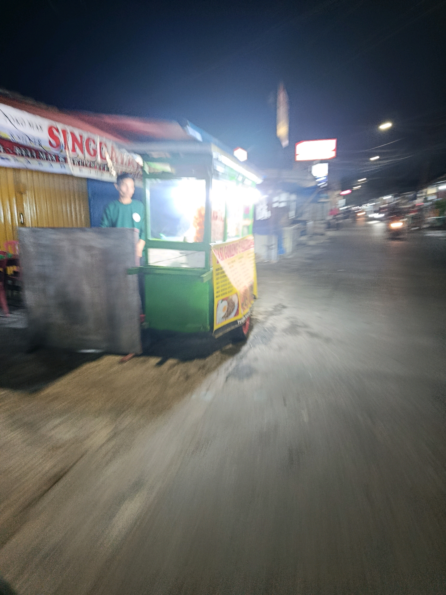 The image captures a street scene at night in Bekasi, Indonesia. In the foreground, a food cart is visible, with a person in a green shirt standing behind it. The cart has a yellow sign with Indonesian text, possibly advertising food items such as "Nasi Goreng" (fried rice). A large, dark, rectangular object, possibly a counter or a display, is positioned to the left of the cart. The background shows a dimly lit street with a few vehicles, including a motorcycle with its headlights on, suggesting ongoing traffic. Several streetlights and illuminated signs contribute to the overall night-time ambiance. The image appears to be taken from a moving perspective, resulting in motion blur, particularly in the road and background elements. The overall scene depicts a typical street-side food vendor operation in a city environment at night.