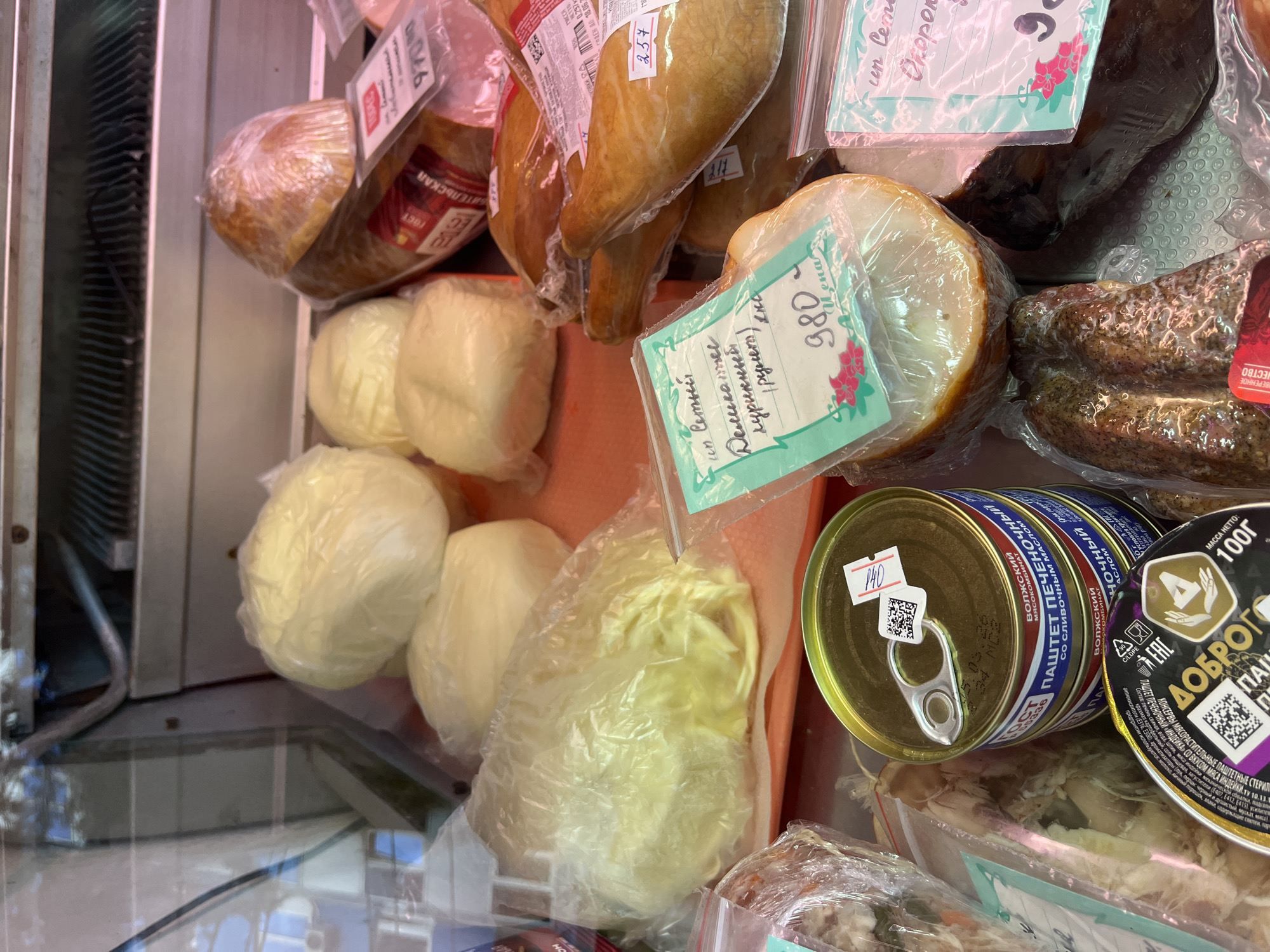This image captures the interior of a refrigerated display case in what appears to be a grocery store or deli in Sochi, Russia. The case is filled with various packaged food items, primarily smoked and dairy products, presented for sale.
In the foreground, several round and pear-shaped items, likely smoked cheese (such as Suluguni), are individually wrapped in clear plastic. Some of these cheese products have price tags attached; one round piece has a handwritten tag reading "980 Цена" (Price 980), and a pear-shaped item shows "257". Above these, more smoked products, possibly meats or fish, are visible in plastic packaging. One larger, darker item is a "Деликатес куриный (рулет)" (Smoked Chicken Delicacy (roll)), as indicated by a handwritten tag which also says "un Сетый". Another tag is partially visible with "un Сем..." and "Окорок" (Ham).
To the right, a stack of two cans of pâté is visible. The top can is labeled "ПАШТЕТ ПЕЧЕНОЧНЫЙ" (Liver Pâté) from "ВОЛЖСКИЙ МЯСОКОМБИНАТ" (Volzhsky Meat Processing Plant) with "со сливочным МАСЛОМ" (with butter). A small price tag on this can reads "140". Another can below it has a best-before date of "05.05.26". Adjacent to these, a round, dark-labeled can of "ДОБРОГО ПАШТЕТ ПЕЧЕНОЧНЫЙ «ИНДЕЙКА»" (Dobrogo Liver Pâté "Turkey") is partially visible, stating "МАССА НЕТТО: 100г" and indicating "ГОДЕН ДО 18.06.2".
The overall scene suggests a commercial setting, with products neatly, though densely, arranged under artificial lighting. Reflections on the glass of the display case are noticeable, indicating the presence of a transparent barrier between the viewer and the food.