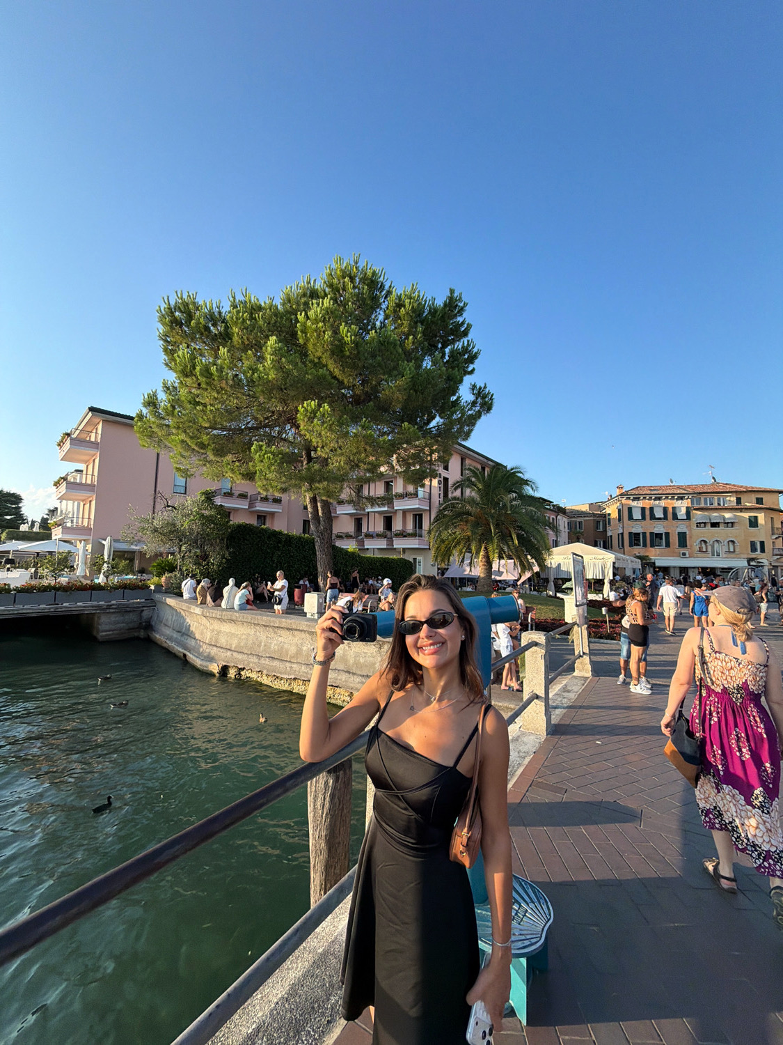 A young Caucasian woman is standing on a paved walkway next to a body of water, holding a camera in her right hand and a cell phone in her left. She is wearing a black spaghetti strap dress, sunglasses, and a light brown purse slung over her shoulder. She has dark brown hair and is smiling at the camera. The walkway is lined with a metal railing on the left and people walking in the background. On the other side of the railing is dark green water with several ducks. Behind the woman are buildings with balconies and trees. The sky is clear and blue. The scene appears to be in Sirmione, Italy, likely near the waterfront, given the presence of the lake and the architectural style of the buildings.