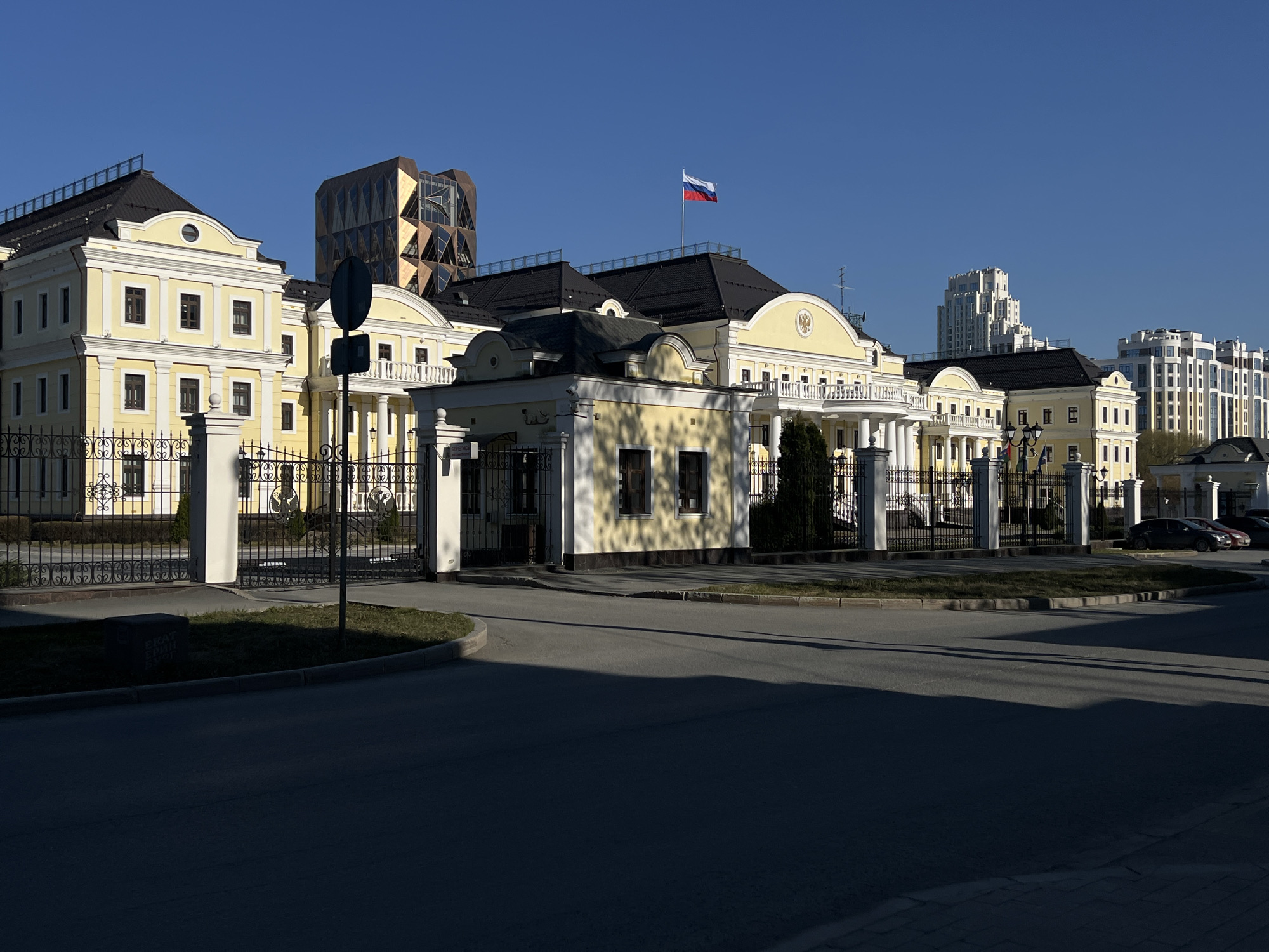 This media file captures a panoramic view of a grand architectural complex on a clear, sunny day in Yekaterinburg, Russia. The primary subject is a large, stately yellow building with white classical columns, balconies, and black sloped roofs, reminiscent of a historical mansion or official residence. A Russian flag flies prominently from a flagpole atop the central section of this building.
In the foreground, a dark asphalt road curves around a grassy median, with long, dark shadows stretching across it, indicating either early morning or late afternoon sunlight. A decorative black metal fence with white pillars encloses the building grounds. A smaller, matching yellow gatehouse stands near the main entrance. Behind the fence, cars are parked on the right side.
In the background, contrasting architectural styles are visible. To the left, a striking modern building with a geometric, faceted facade in shades of brown and gold rises above the yellow complex. Further to the right, a cluster of contemporary white high-rise residential or commercial buildings completes the urban skyline.
The scene is devoid of people, offering a quiet, architectural study. A small, dark stone block on the grass near the foreground is partially obscured by shadow but shows faint Cyrillic text, appearing to read "БЕЛЫЙ ГУСЬ" (White Goose). The overall impression is one of formal grandeur set within a modern city environment.