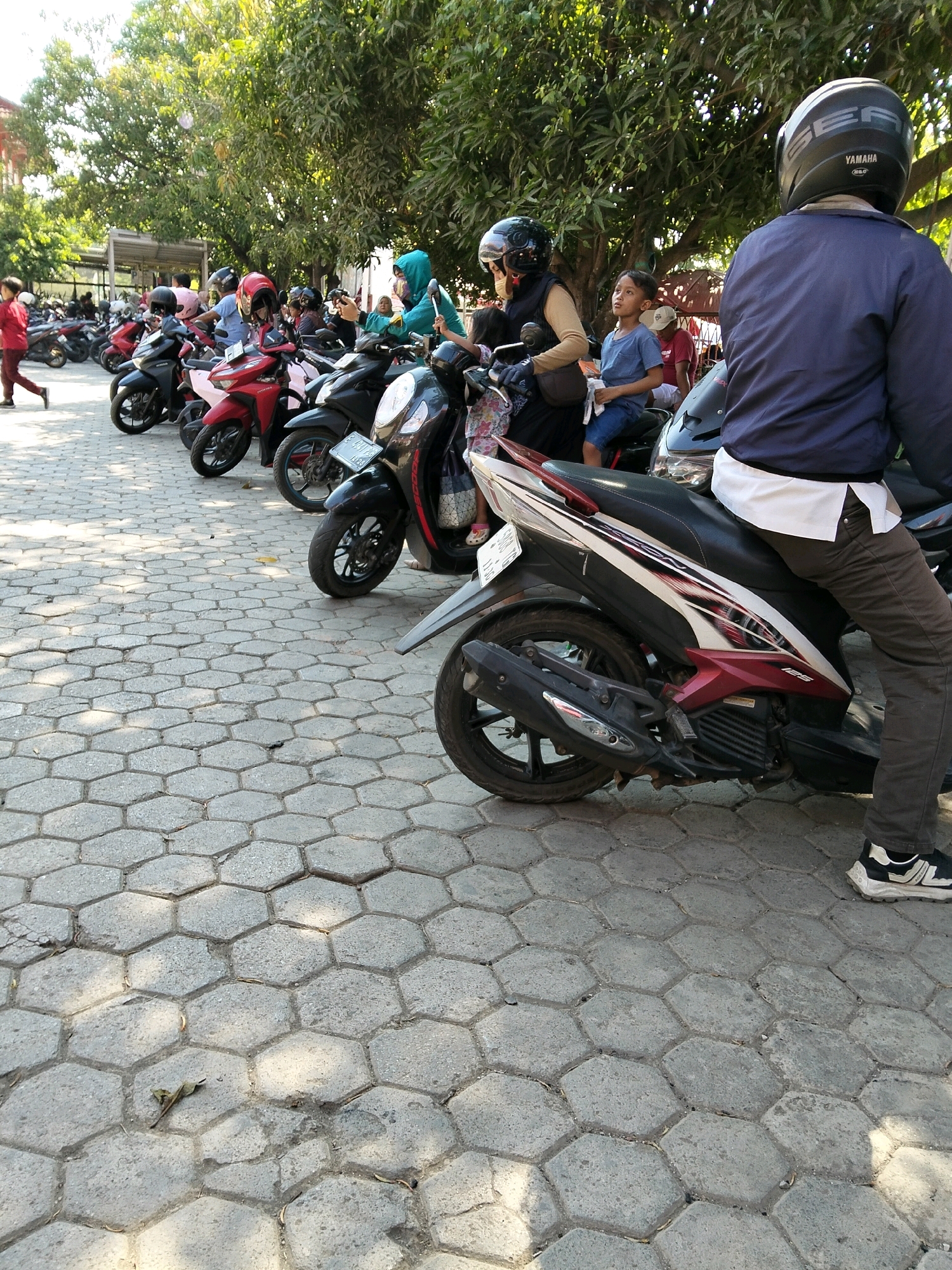 In Indramayu, Indonesia, a parking lot paved with hexagonal interlocking concrete blocks is filled with parked motorbikes. A woman wearing a hijab and a blue jacket, her face obscured by a dark helmet, sits on a motorbike in the foreground. To her left, a young boy in a blue t-shirt and shorts looks up, holding a piece of white fabric. Behind them, a line of motorbikes extends into the background. To the left of the frame, a person in a red shirt walks away from the viewer. Trees with lush green foliage provide shade over the scene.
