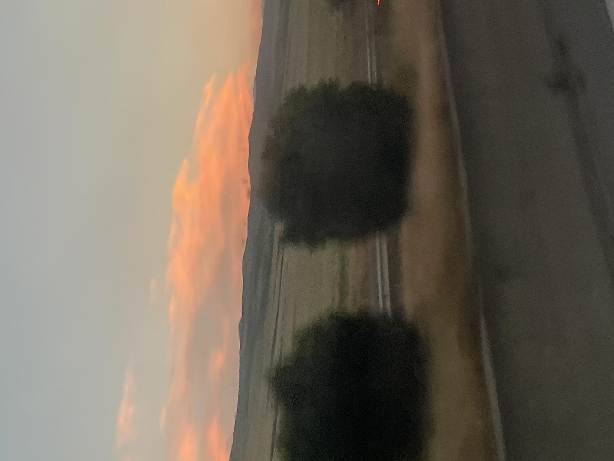 A blurred, horizontal, eye-level shot depicts a landscape at dusk. The sky is a gradient of soft blue and peach, with wispy, orange-tinged clouds. Below the sky are rolling hills covered in green fields. In the foreground, two dark, bushy trees obscure a portion of the landscape. A highway is visible between the trees, with a few distant lights suggesting vehicles. The lower part of the image is dominated by a dark, blurred foreground that appears to be a road surface. The overall impression is one of a serene, natural setting at the end of the day. The image does not contain any recognizable landmarks or features that would allow for specific location identification within the city of Dzveli Kanda, Georgia.