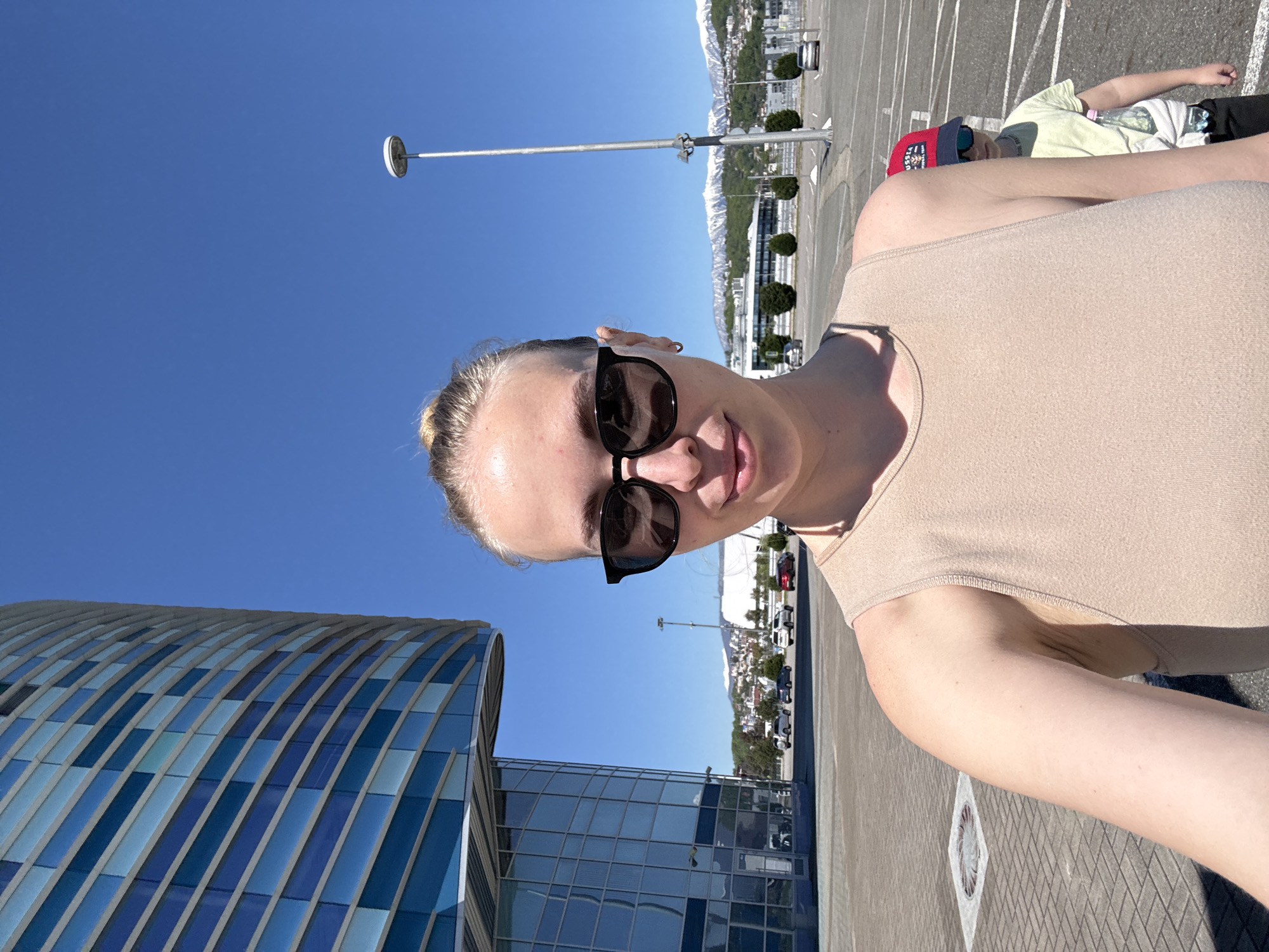This selfie captures a woman on a clear, sunny day in Sochi, Russia, with distinct snow-capped mountains in the background. The primary subject is a fair-skinned woman with blonde hair pulled back, wearing black sunglasses and a light-colored sleeveless top. She is looking directly at the camera with a slight smile, suggesting a relaxed mood.
To her right, a second person is partially visible, wearing a light-colored t-shirt, dark pants, and a red baseball cap, appearing to be resting or sitting in the paved area. A water bottle is visible next to them.
On the left side of the frame, a striking modern building with a curved, blue and white striped glass facade dominates the foreground. The scene is set in a large, open paved area, likely a parking lot, with white markings visible on the ground. Tall street lights are interspersed throughout this area.
The weather is bright and clear, indicated by the vibrant blue sky with no clouds. The distant snow-capped mountains confirm the mountainous geography of the region, aligning with the location context of Sochi. No discernible text is visible on any objects or people.
