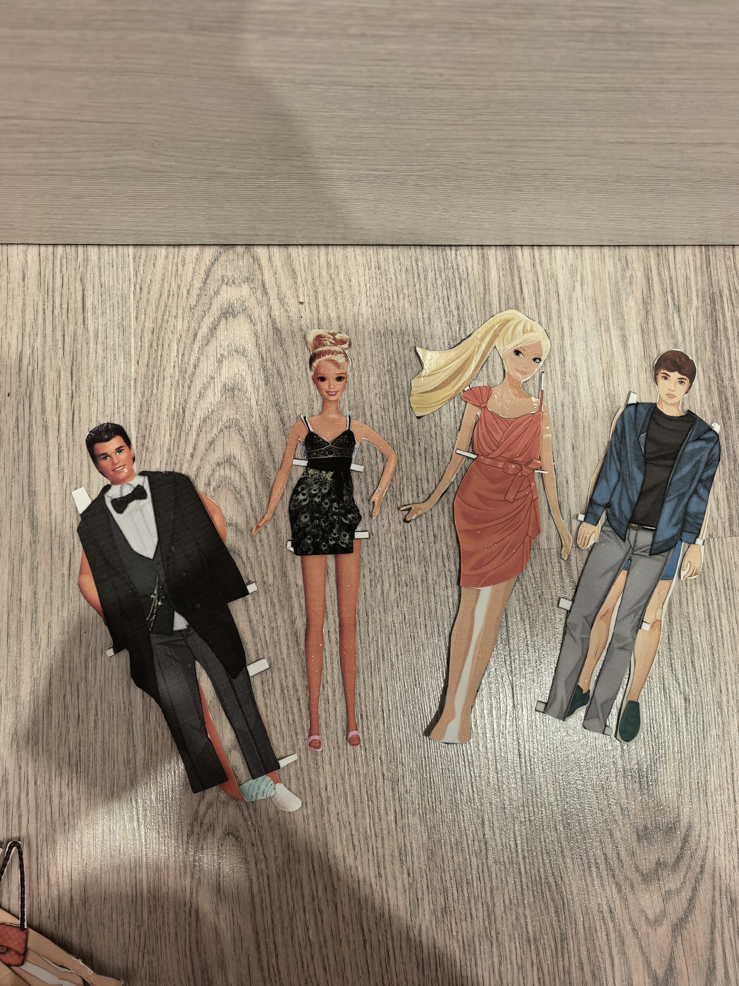 Four paper doll cutouts lie on a wood-grain surface, likely flooring or a tabletop. From left to right, the dolls are: a man in a tuxedo; a woman in a black dress; a woman in a red dress; and a man in jeans and a jacket. The dolls all have tabs on the sides for attaching clothing. The scene appears to be indoors, with simple lighting and no other visible objects except for a small portion of a shopping bag at the lower left corner. There is no information to suggest this image was taken in Sterlitamak, Russia.