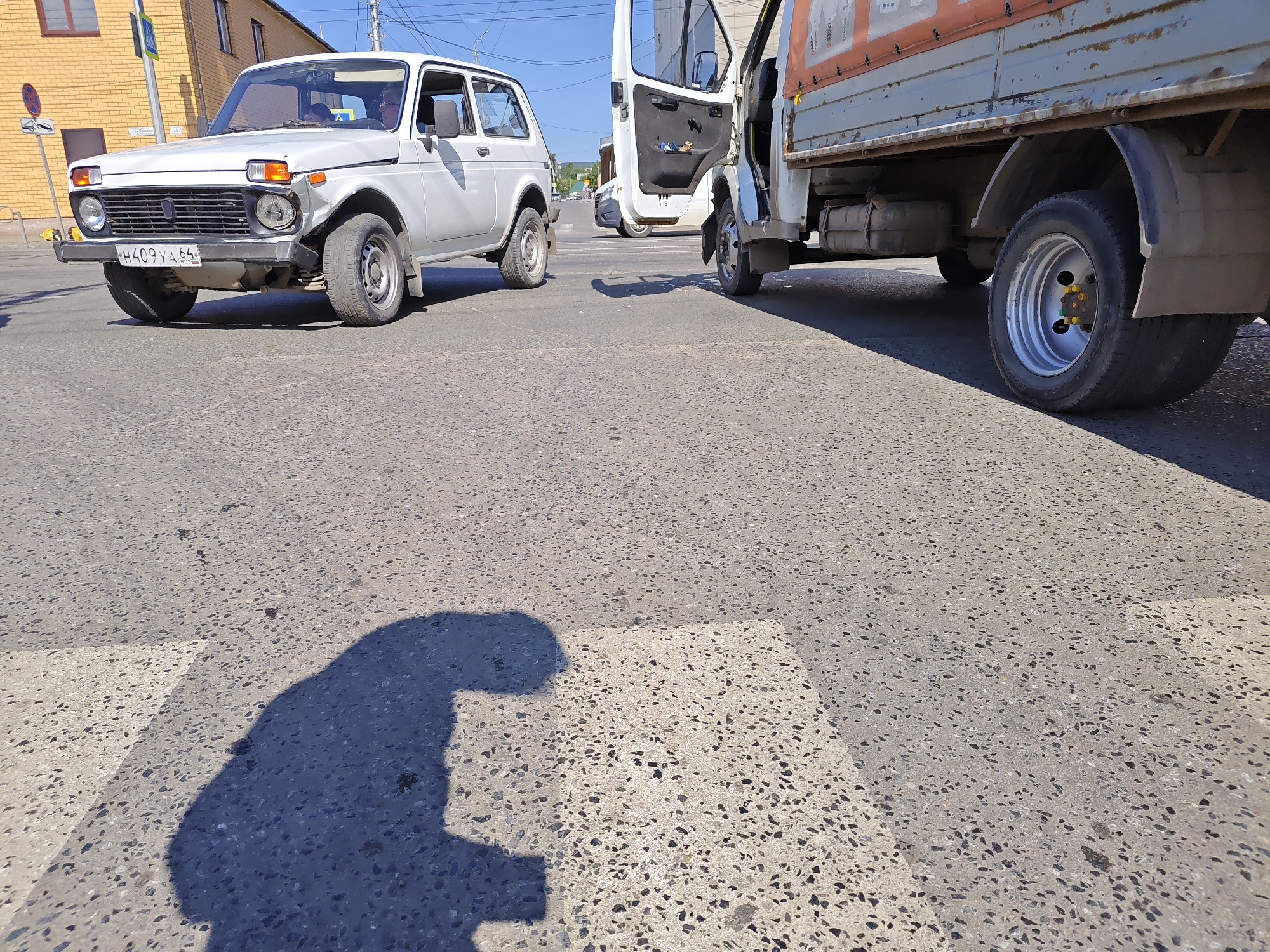 The image shows a street scene with two vehicles, a white car and a light-colored truck, on a paved road. The white car, a Lada Niva, is on the left side of the frame, facing the viewer. It has a license plate that reads "H409YA 64 RUS". The front of the car appears to have some damage. The truck is on the right side of the frame, with its driver's side door open. The truck is facing away from the viewer. The ground is a crosswalk with white stripes.
In the foreground, a shadow of a person is cast on the crosswalk. The person appears to be holding something, possibly a phone. The shadow suggests the sun is to the left and slightly behind the viewer, indicating it is likely daytime. The sky in the background is blue and clear, suggesting fair weather.
Behind the vehicles, buildings are visible, including a yellow brick building on the left. There are also utility poles and wires. The overall scene depicts a moment at a street intersection. The presence of vehicles and a crosswalk suggests an urban or suburban environment. The lighting and clear sky suggest the photo was taken on a sunny day.