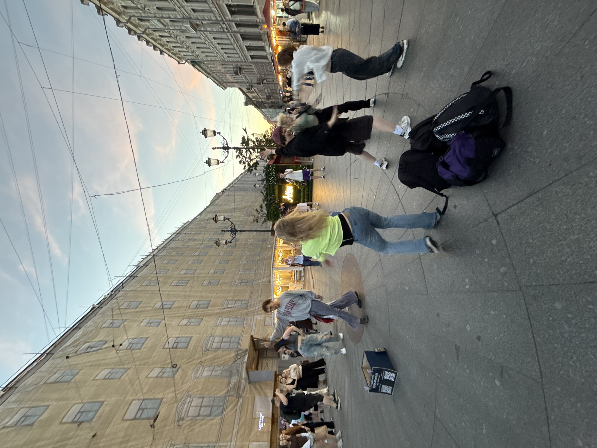 The image captures a street scene in Saint Petersburg, Russia, likely during the early evening hours. A group of young people are engaged in dancing in an open pedestrian area. Several buildings line the street; one building on the left is covered in a large beige tarp, possibly undergoing renovation, and displays numerous faux windows. The building on the right features more traditional architecture with visible windows and ornamentation. Streetlights with multiple lamps are visible, along with overhead wires crisscrossing the sky, which is beginning to show hues of pink and orange from the setting sun.
In the foreground, a collection of bags, including a purple backpack and black duffel bags, are placed on the ground, suggesting the dancers may have arrived with their belongings. A blue box, potentially a donation box or prop, is also present on the pavement with QR codes and text printed on it.
The dancers themselves are casually dressed in jeans, t-shirts, and sweatshirts, exhibiting dynamic poses and movements. The crowd of onlookers is scattered around the periphery, observing the performance. The ground is paved with large, grey stone tiles, arranged in a circular pattern in the area where the dancing is taking place. The overall atmosphere appears lively and informal.