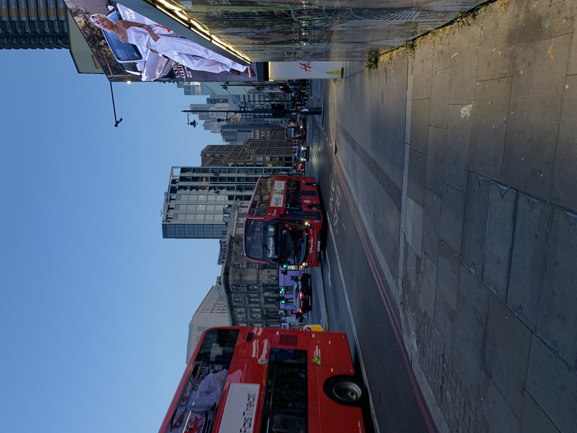 The image captures a vibrant street scene in London, United Kingdom, during what appears to be late afternoon or early evening under a clear blue sky. The main subjects are two iconic red double-decker buses, characteristic of the city, which are part of the traffic flow on a multi-lane road.
The street is lined with a mix of modern, glass-fronted buildings and older, more traditional urban architecture. On the left, a large digital billboard displays a fashion advertisement featuring a woman in a white outfit next to a car. Visible text on the billboard includes "S/S 2026 COLLECTION" and "FASHION." Below the billboard, an H&M logo is clearly seen.
One of the double-decker buses in the midground displays the route number "8" and the destination "Bow Church." The bus in the foreground shows the number "11," with partial text like "TEST" also visible. Several other cars are present in the traffic. The pavement on the right side of the road is constructed from large, grey square tiles. Road markings, including a partially visible "BUS LANE" designation, delineate the traffic areas. The scene portrays a typical busy city environment, bustling with vehicles.