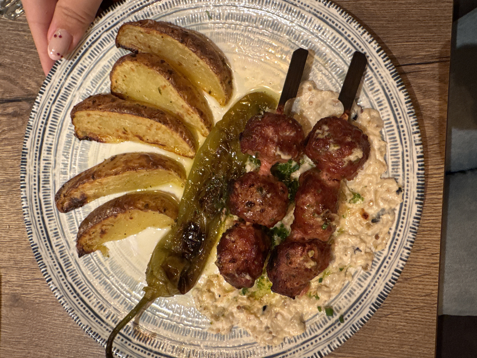 A plate of food is presented on a wooden table. The plate has a blue and white patterned rim and contains roasted potato wedges, a roasted green pepper, and meatballs on skewers. The meatballs are served on a creamy sauce, possibly hummus or tahini, with chopped herbs. A person's hand with manicured nails is visible on the right side of the image, reaching towards the plate. The image was taken in the city of Varna, Bulgaria.