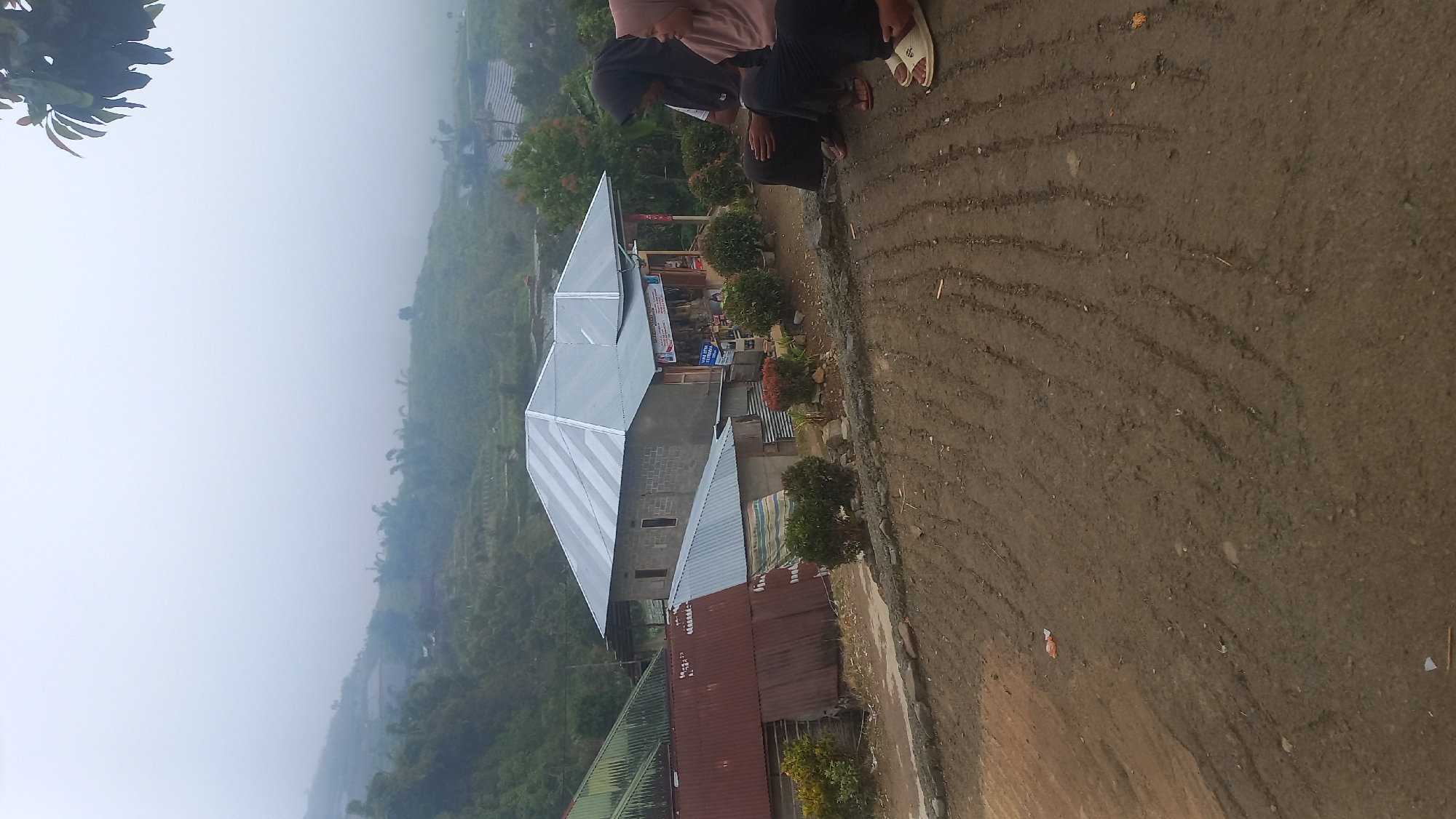 The image depicts a rural hillside scene in Indonesia, possibly a village in a mountainous region, obscured by mist. In the foreground, a muddy path with tire tracks leads towards a residential area. Two women, both wearing hijabs, are seated on the edge of this path, facing towards the right of the frame. One woman, on the left, is wearing a black hijab and dark clothing. The woman on the right wears a beige hijab and also dark clothing. Behind them, a cluster of houses is visible, including a two-story building with a silver, pitched roof, constructed from grey brick. Adjacent to it is a smaller building with a corrugated tin roof, partially rusted and red. In front of the brick building, a small shop or market stall is set up with goods displayed. The surrounding landscape is hilly and covered in lush greenery, with terraced fields visible in the distance, further emphasizing the rural setting. The overall atmosphere is serene and somewhat diffused due to the mist.
