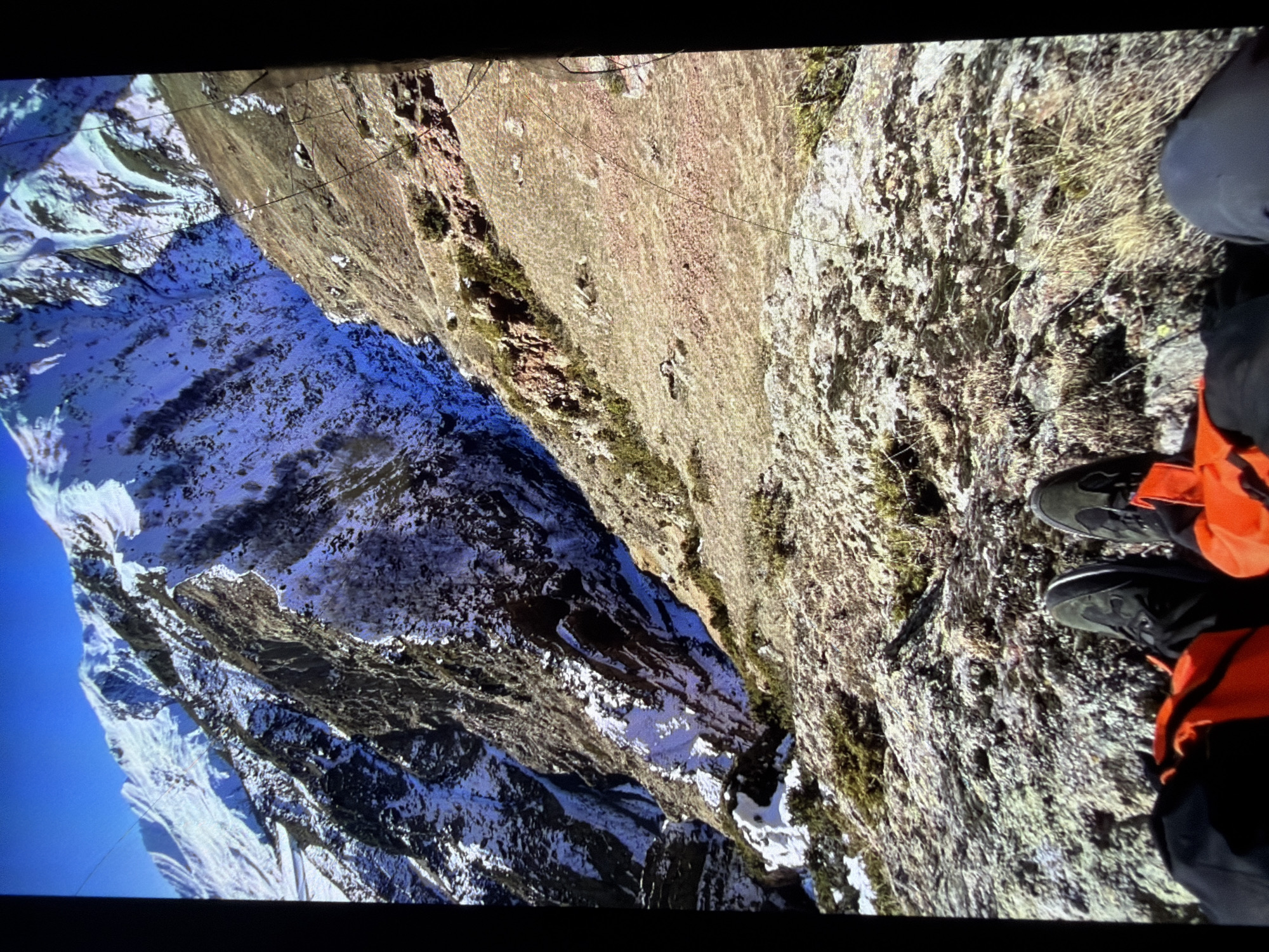 The image captures a first-person perspective from a rocky outcrop overlooking a steep, snow-dusted mountain slope. In the foreground, a person's legs are visible, wearing dark hiking boots and possibly bright orange outerwear, suggesting they are geared for outdoor activity. The mountain scenery dominates the frame, with rugged terrain, patches of snow, and rocky formations. The sky is a clear, vibrant blue. The overall impression is one of a remote and mountainous landscape, likely a hiking or climbing location.