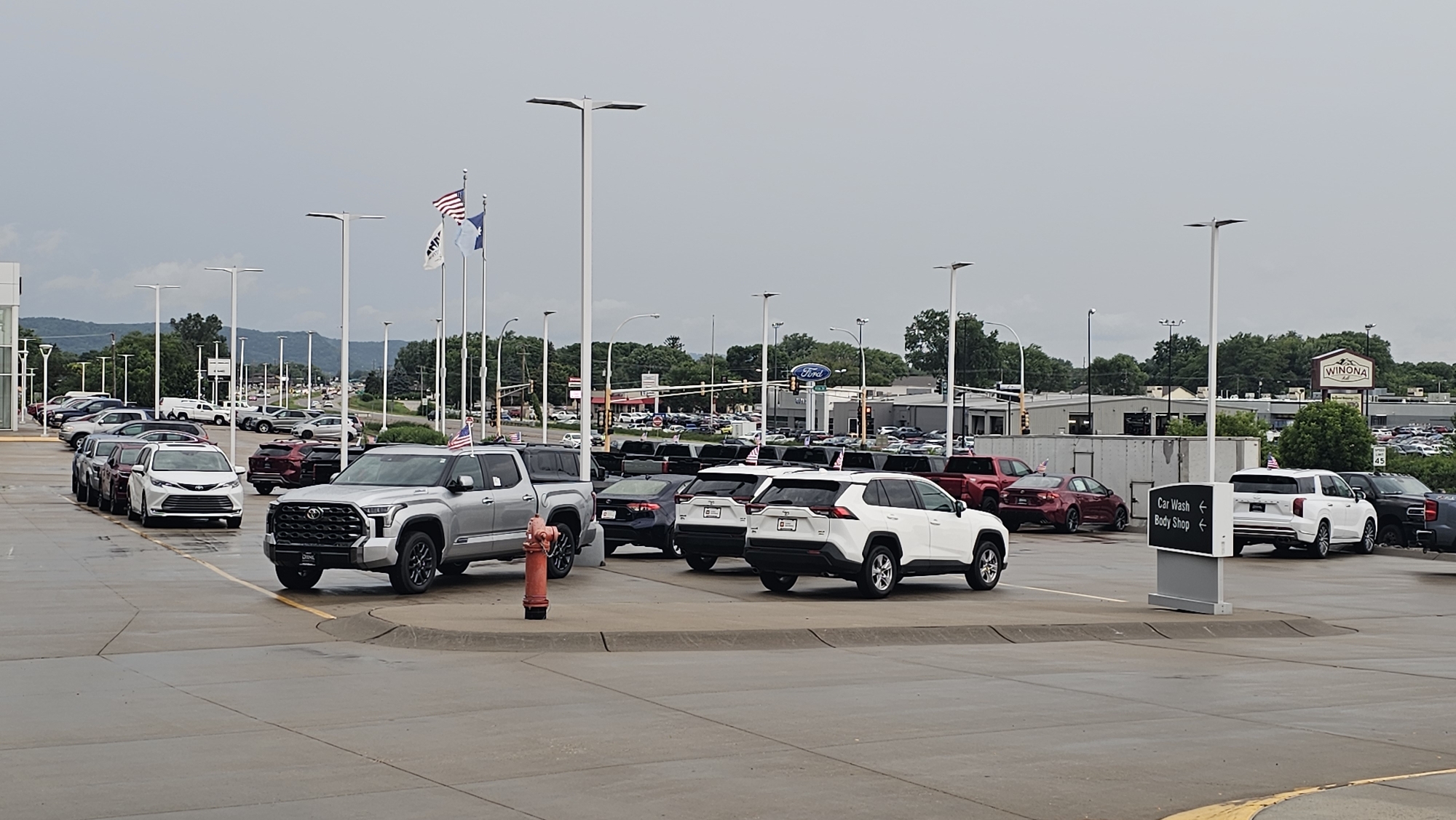 The image shows an outdoor car dealership in Winona, United States of America. Numerous cars, predominantly SUVs and trucks, are parked in rows on a wet asphalt lot. In the foreground, a silver Toyota Tundra pickup truck is parked, with a red fire hydrant next to it. To the right, a white Toyota RAV4 SUV and a white Toyota Highlander SUV are visible. Further back, other vehicles are parked, including sedans and more SUVs. In the background, a sign for "Winona" with a logo is visible on a building, along with a Ford dealership sign. The sky is overcast, and there are trees and distant hills in the background. Flags, including the American flag, are flown on tall poles.