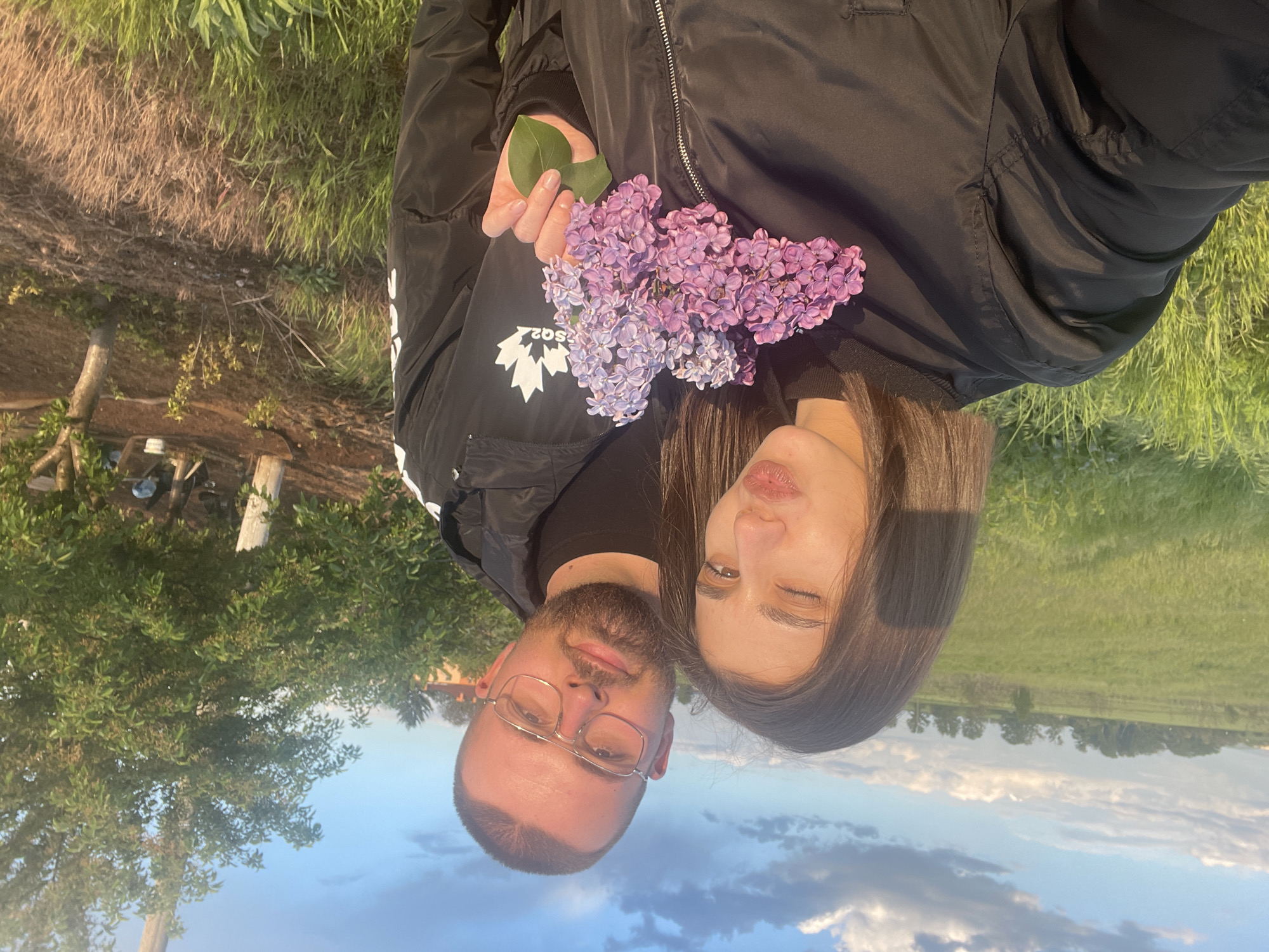 A man and a woman are standing outdoors, posing for a photograph. The man, who has a beard and wears glasses, is on the left. The woman, with long dark hair, is on the right. Both are wearing dark-colored jackets. Between them, the woman is holding a bouquet of lilac flowers, and the man is holding a single green leaf. The background shows a green grassy area, some trees, and a blue sky with clouds. While the information necessary to identify the specific location within Sofia, Bulgaria is not visible in this picture, the setting appears to be in a park or a rural area.