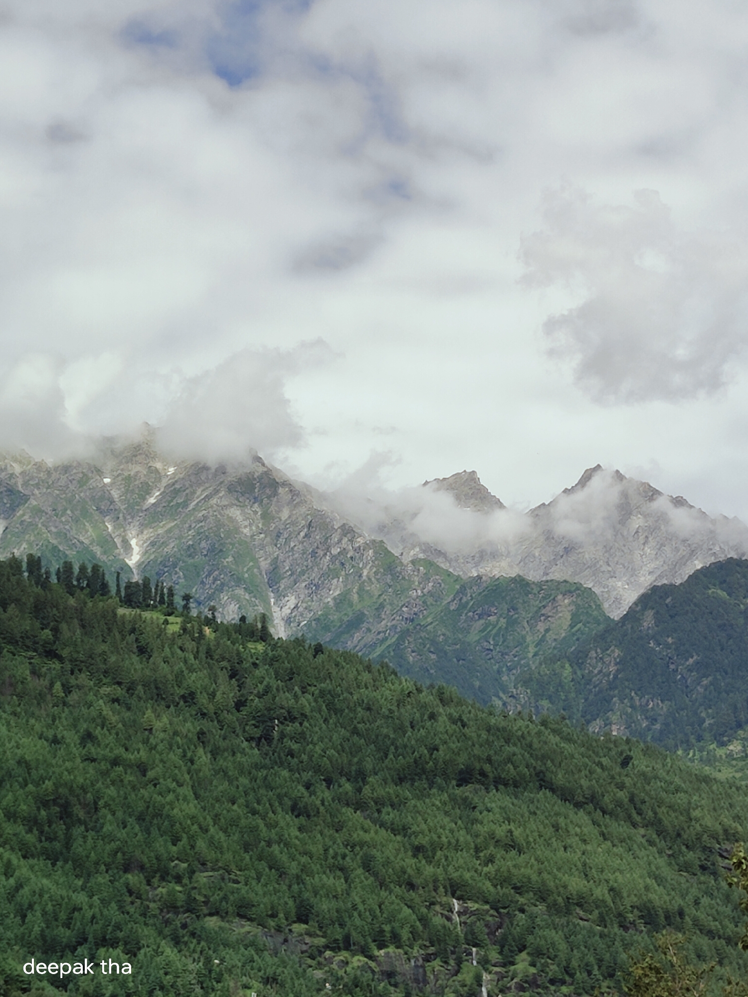 The image depicts a mountainous landscape in Vashisht, India, characterized by lush green forests covering the lower slopes and rugged, rocky peaks dominating the background. The sky is overcast with scattered clouds, some of which partially obscure the mountain summits, lending a dramatic and somewhat misty atmosphere to the scene. A dense forest of coniferous trees blankets the mid-ground, with individual trees clearly visible and creating a textured green carpet. In the distance, the mountains exhibit sharp, jagged contours, with patches of snow or lighter colored rock visible on their upper reaches. The overall impression is one of natural grandeur and a remote, possibly cool, mountainous environment. The text "deepak tha" is visible in the lower left corner of the image.