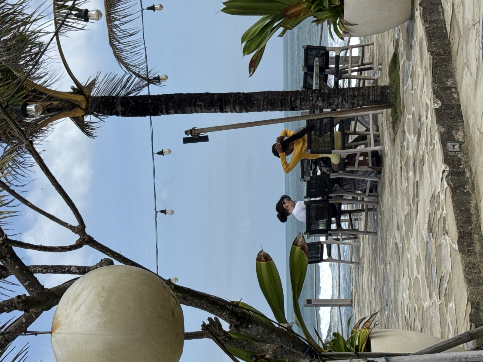 A medium shot captures an outdoor seating area by the ocean in Mengwi, Indonesia. The scene features two individuals seated at tables with chairs, facing away from the camera, toward the ocean. One person, wearing a white top, is seated on the left, and another, wearing a yellow top, is on the right, with their back to the viewer. The environment includes a stone-paved patio, a railing overlooking the water, and palm trees with hanging lights. Large potted plants are positioned on either side of the patio. The ocean is visible with moderate waves under a cloudy blue sky. A large, spherical decorative object hangs from a tree branch in the foreground.