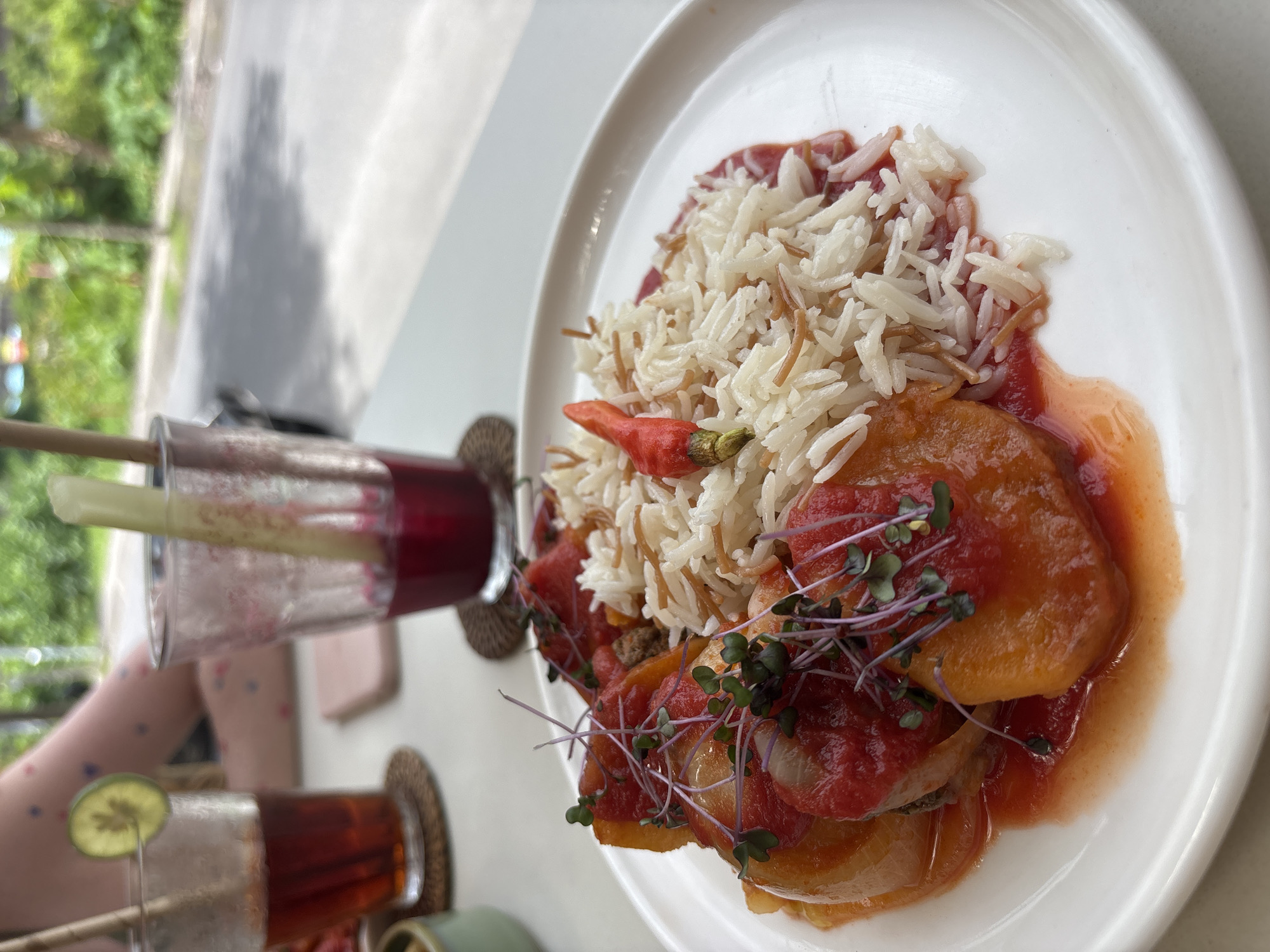 The image shows a plate of food and two drinks on a white table. The food appears to be rice with vegetables in a tomato sauce. The rice is a mix of white rice and what appears to be vermicelli. The vegetables seem to be squash or potatoes. The drink on the left is a dark red color and has a straw in it. The drink on the bottom is also dark, with a green garnish and straw. The scene is outdoors with trees in the background. The location is in Mengwi, Indonesia, based on the image context.