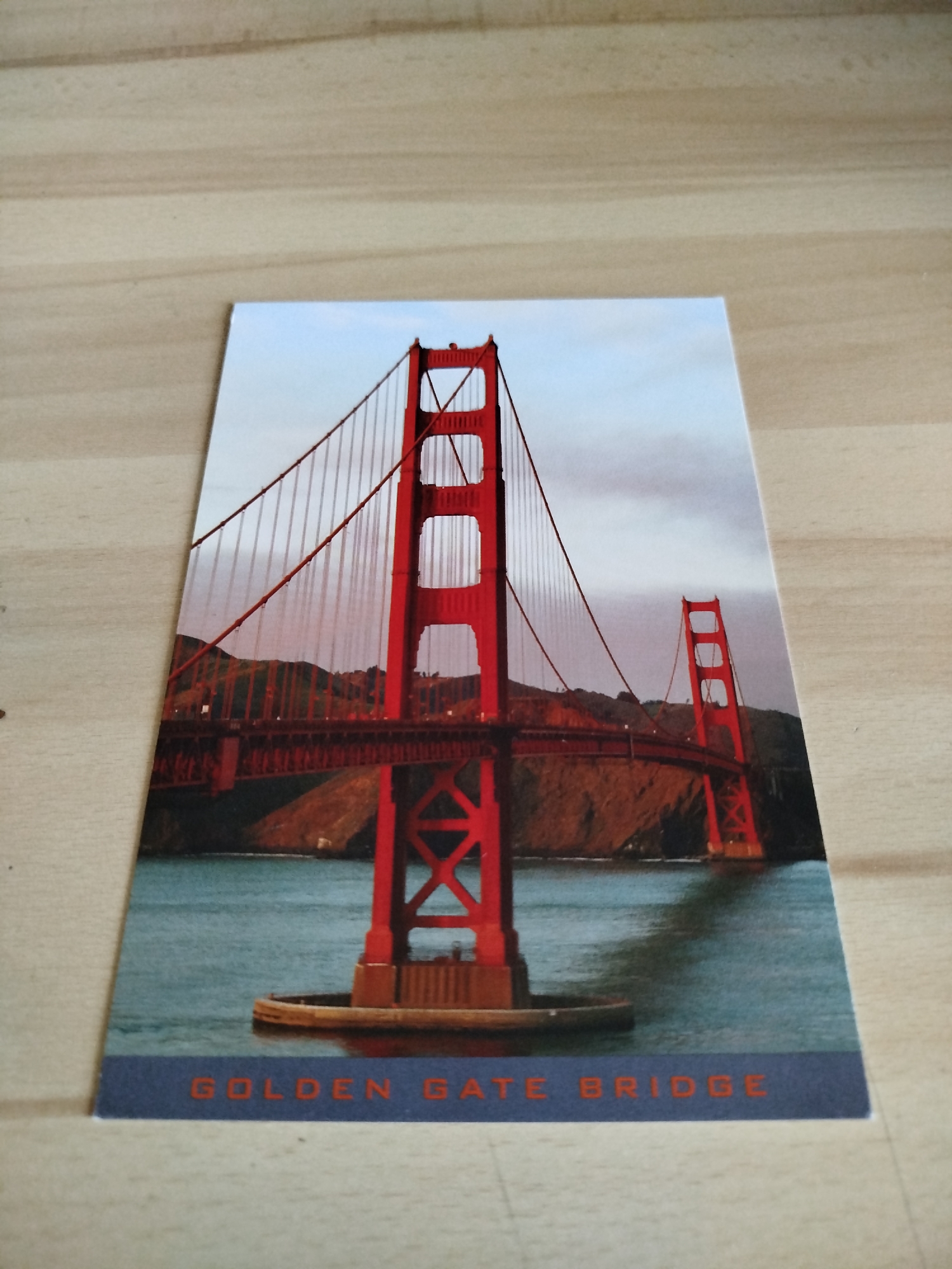 The image is a postcard depicting the Golden Gate Bridge in San Francisco, California. The bridge is red and spans a body of water. The text "GOLDEN GATE BRIDGE" is printed at the bottom of the card. The postcard is lying on a light brown wooden surface. The city of Varna, Bulgaria is not represented in this image.