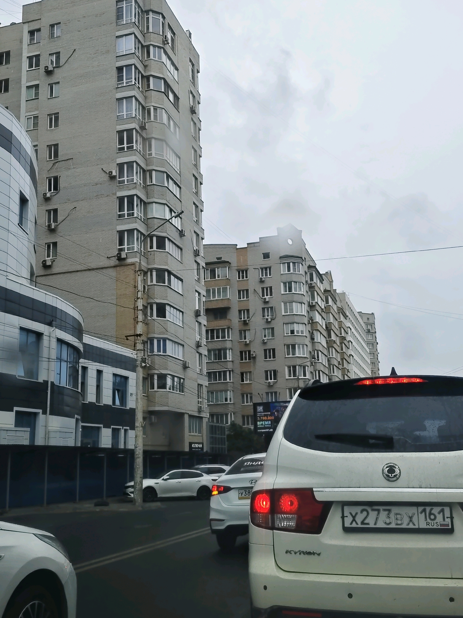 The image depicts a street scene in Rostov-on-Don, Russia, with multiple apartment buildings and cars. The weather appears overcast. In the foreground, the rear of a white SUV, identified by the "KYRON" badging and a license plate reading "X273BX 161 RUS," is visible. Red brake lights are illuminated. To the left of the SUV, another white car is partially visible. In the middle ground, several white sedans are in traffic. The background features several tall, multi-story apartment buildings constructed of grey brick, some with balconies and numerous windows. A modern, curved building with dark and white sections is also present on the left side of the image. Utility poles and wires are visible along the street.