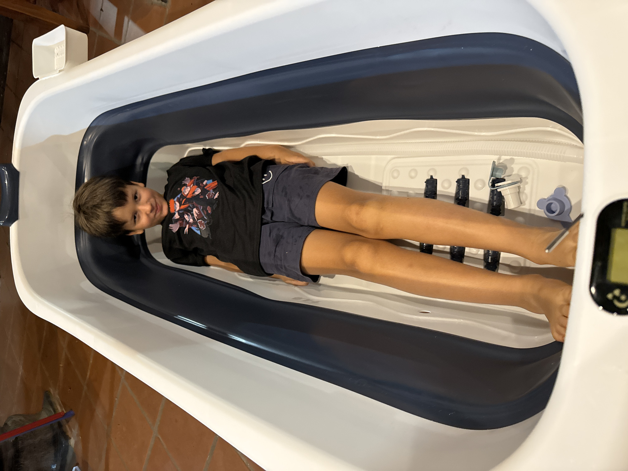 A child lies in a hydrotherapy tub. The tub is white with a dark blue rim. The child is wearing a black t-shirt and dark shorts. The tub is in a room with a tiled floor. The location is Mũi Né, Vietnam.