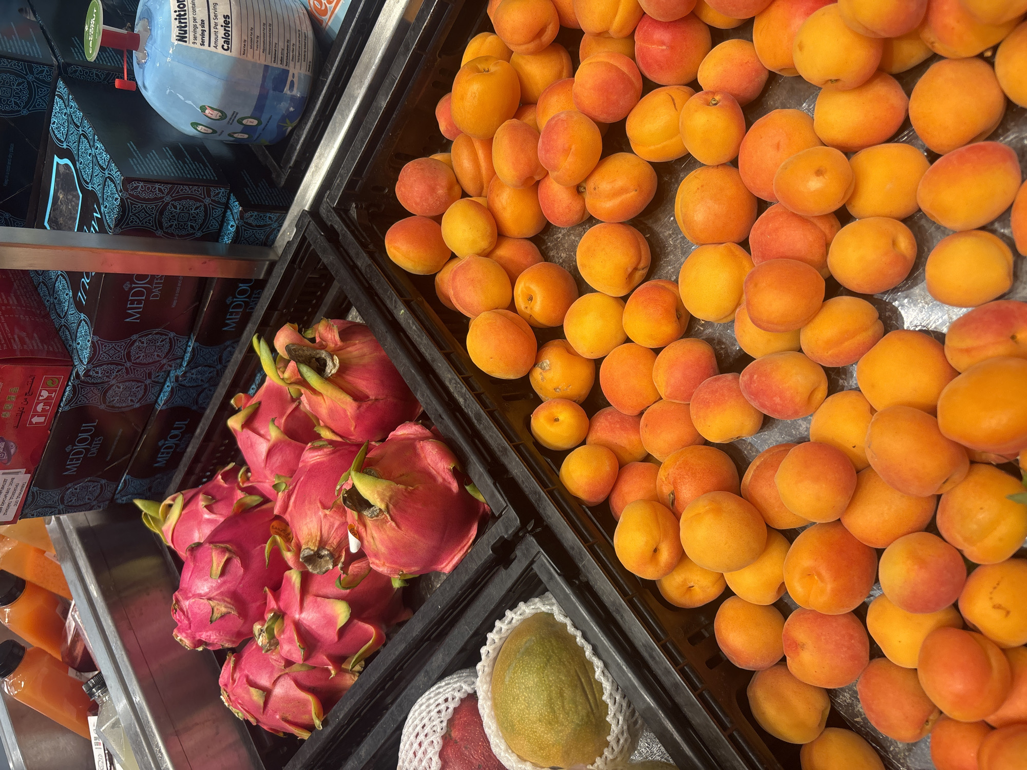 The image shows two black plastic bins of fruit on a grocery store shelf. The bin on the right is filled with apricots, while the bin on the left contains dragon fruit and a single mango wrapped in protective netting. Above the bins is a shelf containing containers of juice, boxes of Medjool dates, and a blue bottle labeled "Nutrition." The labels on the date boxes include "Keep in a cool and dry place" and the text includes Cyrillic and Thai characters, along with Arabic and English, suggesting the products are imported. The "Nutrition" bottle's label includes "Serving size" and "Calories." The location of the scene is not provided in the image, and I cannot determine it.