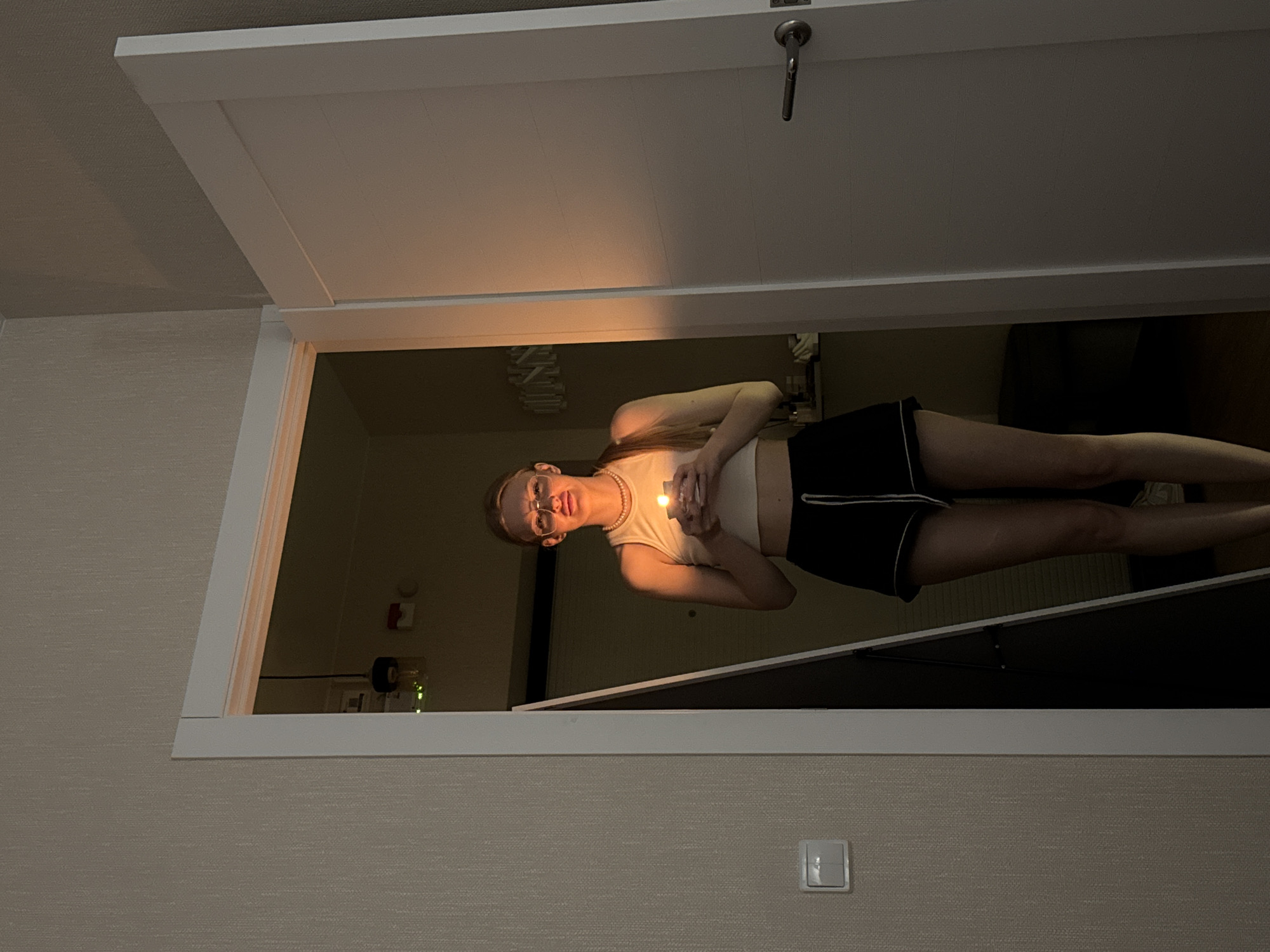 The image depicts a young woman standing in an open white doorway, looking directly at the viewer. She is illuminated by a small, glowing light source, possibly a candle or small torch, which she holds clasped in both hands near her chest. She has light-colored hair, wears glasses, a light-colored crop top, and dark shorts with a white trim. A light-colored beaded necklace is visible around her neck. Her expression is calm and direct.
The doorway opens into a dimly lit interior space. Faint details in the background include what appears to be a light fixture on a wall and a power outlet or device with a green indicator light on an adjacent wall. The walls immediately surrounding the doorway are covered in a light-colored, textured wallpaper. A white light switch is located on the wall in the lower right portion of the image. The white door itself is slightly ajar, with a metal handle visible on its right side.
The primary light source in the scene is the small glow held by the woman, suggesting it is either nighttime or a very dark indoor environment. The overall atmosphere is quiet and subdued. No specific weather conditions are discernible from the indoor setting. The scene appears to be a modern residential interior, consistent with a location like Perm, Russia. There is no visible text in the image.