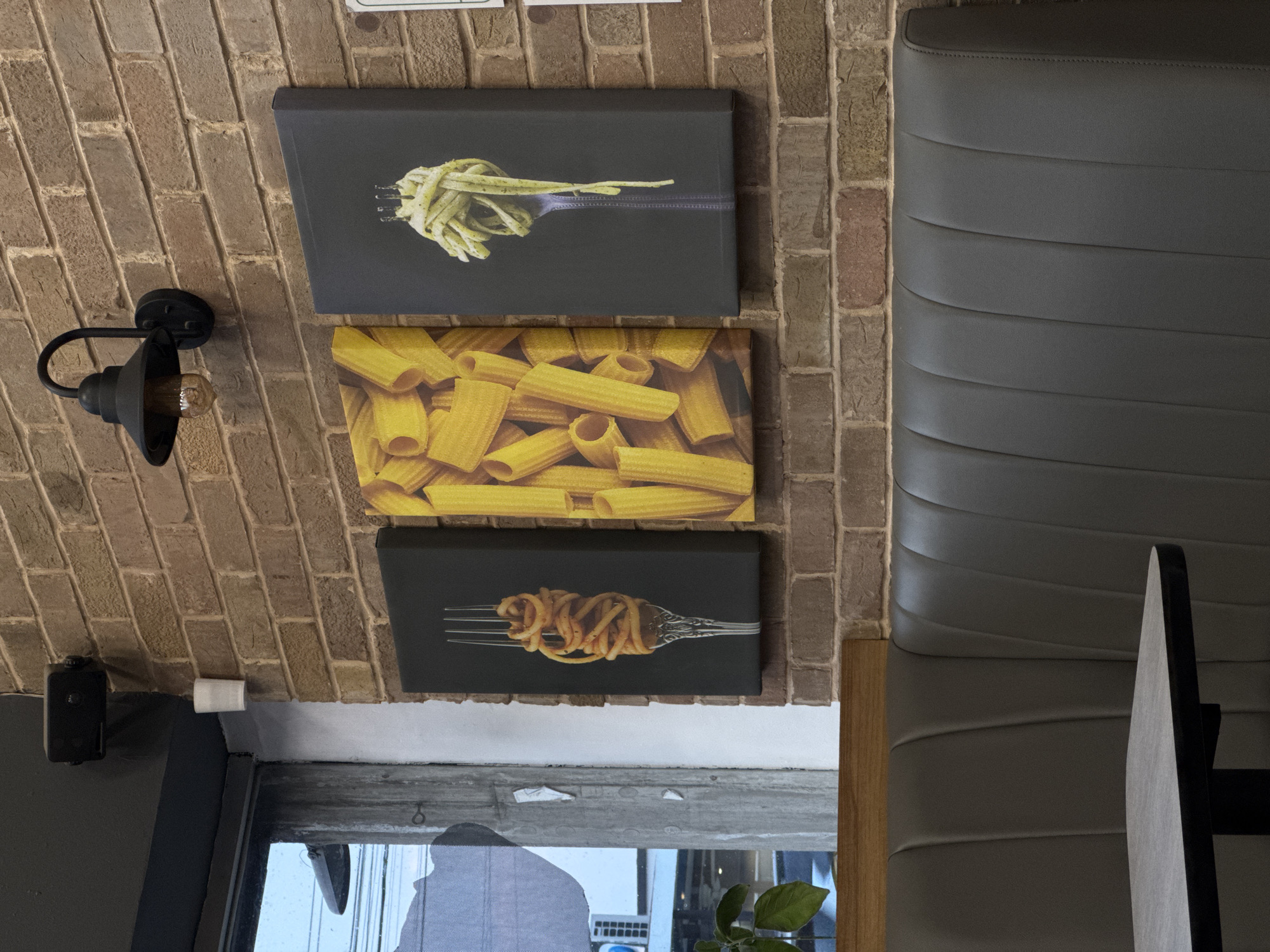 The scene depicts a restaurant or cafe interior with a rustic aesthetic. A brick wall serves as a backdrop for three framed artworks featuring different types of pasta: spaghetti with pesto, rigatoni, and spaghetti with tomato sauce. A black, industrial-style wall lamp is mounted on the brick wall to the left of the artworks. A padded, grey banquette runs along the right side of the image, indicating a seating area. A partial view of a window reveals a glimpse of the outside, showcasing urban elements and possibly vegetation. Based on the information available, it is not possible to confirm the location of the cafe is in San Andrés Cholula, Mexico.